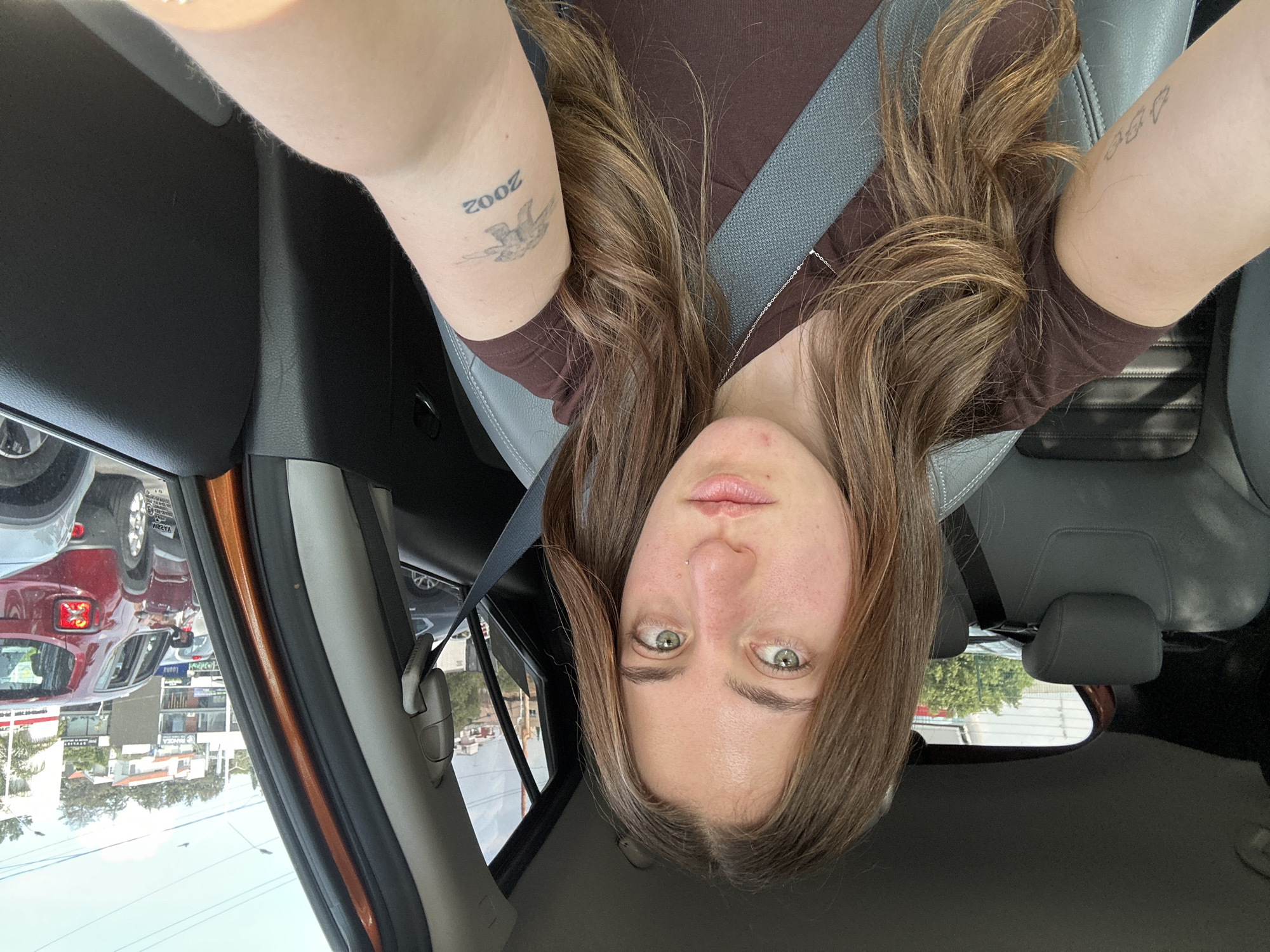 A young woman with brown hair and green eyes is in the driver's seat of a car, wearing a brown t-shirt and a seatbelt. The car is parked on a street in Huixquilucan de Degollado, Mexico. Tattoos are visible on both of her arms. Other cars and buildings are visible through the windows.