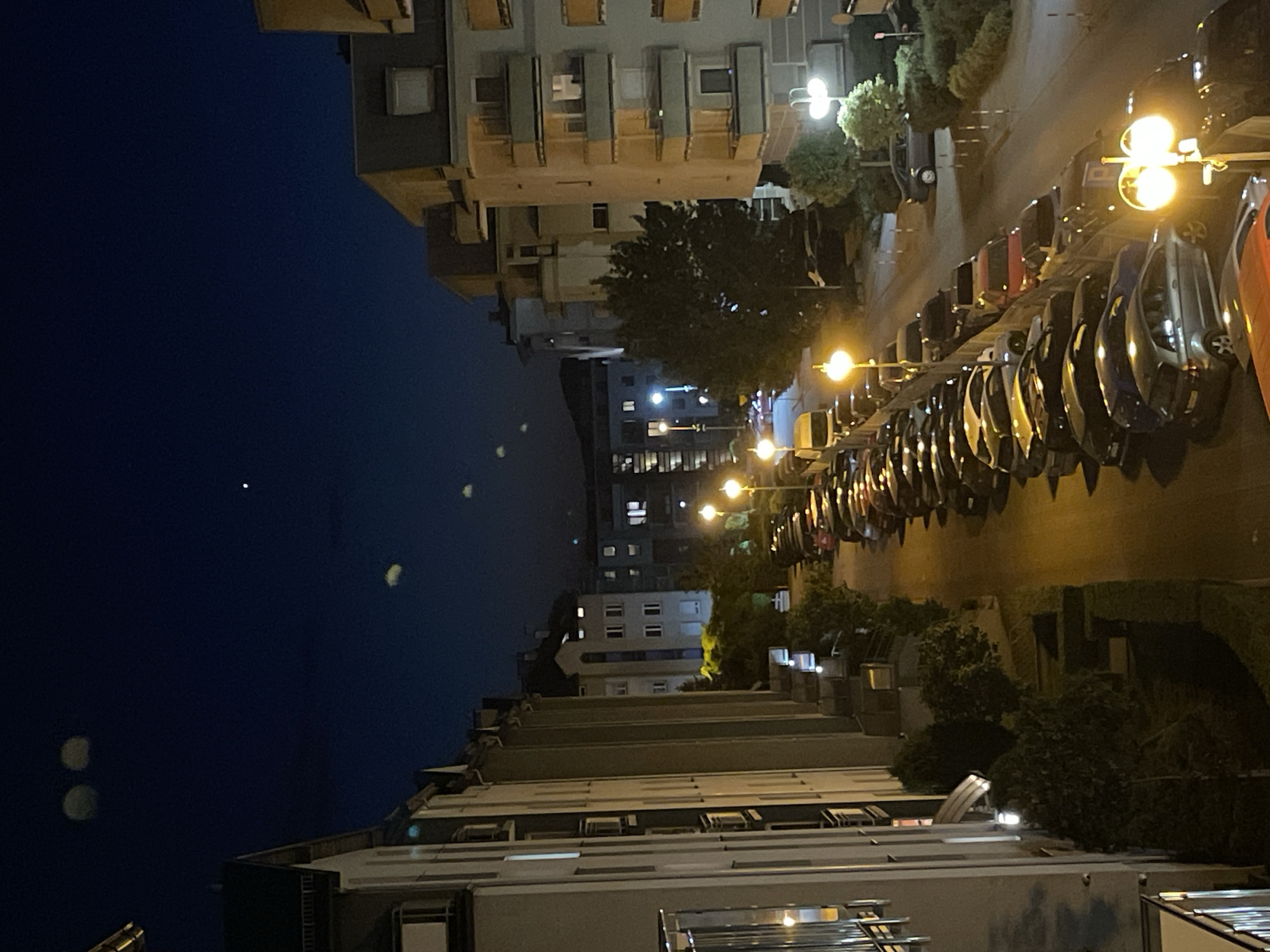 The image depicts an urban scene at night. A line of parked cars occupies the right side of the frame, illuminated by streetlights. Buildings of varying heights are visible on both sides of the street. The sky is dark blue, and several blurred spots are present, possibly artifacts or light sources. No people are visible. The location appears to be a residential area.