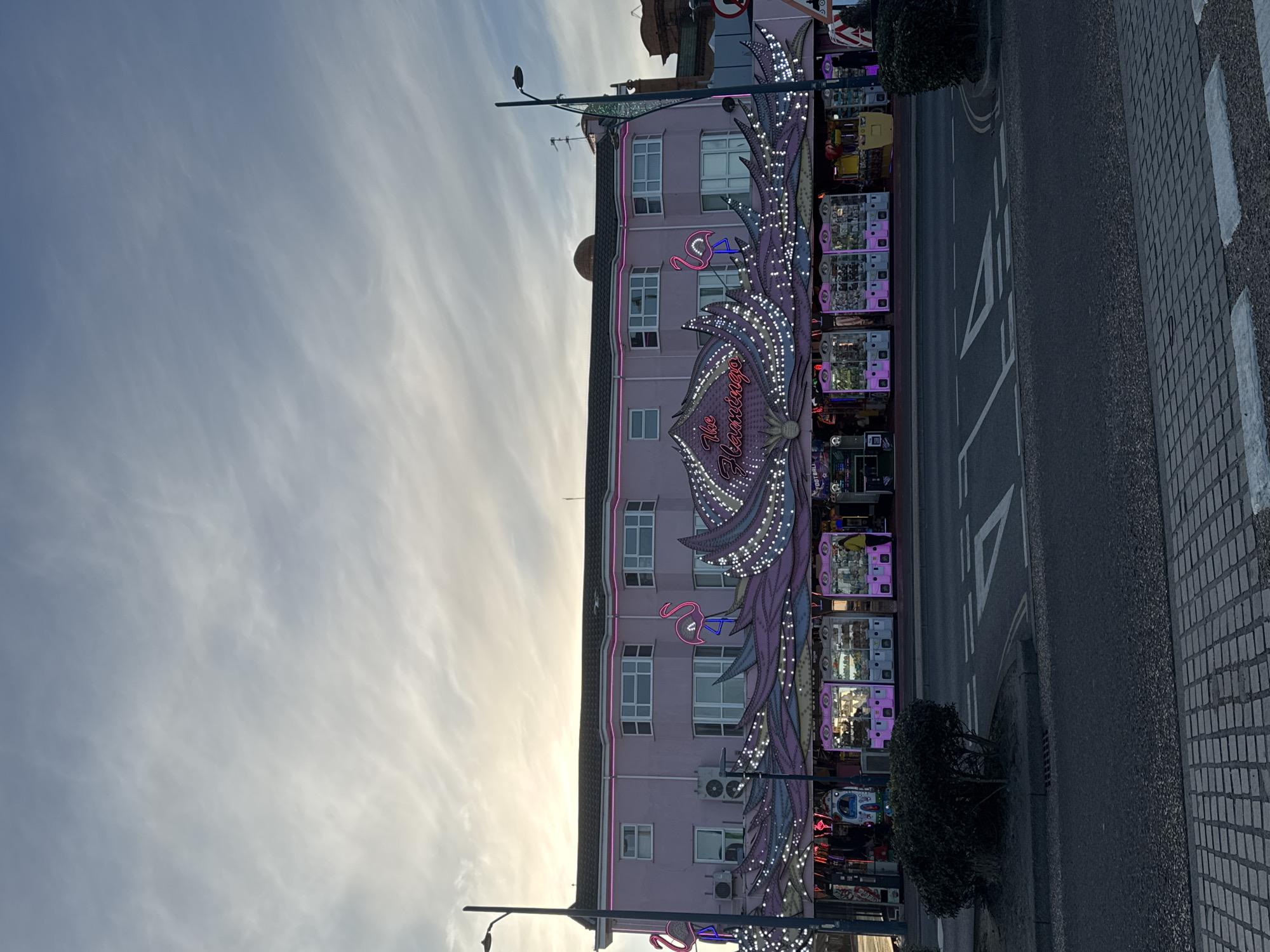The image depicts a building called "The Flamingo" in Gorleston-on-Sea, United Kingdom. The building is a pink, multi-story structure with a large, decorative, illuminated sign that reads "The Flamingo" and features flamingo motifs. The sign is adorned with numerous lights. The building appears to house some form of arcade or entertainment center, with rows of game machines visible through the windows. A street runs adjacent to the building, marked with white road markings. Streetlights are visible along the street. The sky is visible in the background and is partly cloudy.