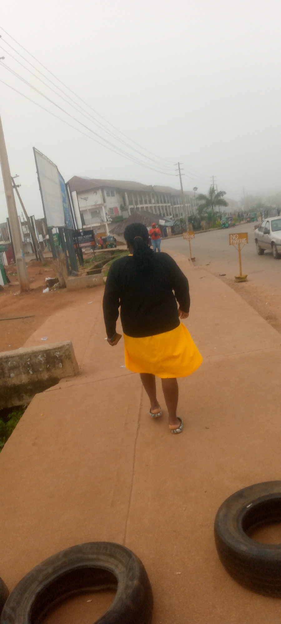 A full shot from behind shows a Black woman wearing a black long-sleeved shirt and a bright yellow skirt walking away from the camera on a concrete sidewalk. She is wearing flip-flops. The environment appears to be a city street, possibly in Gura Top, Nigeria, with a road visible to her right. Buildings, including a large multi-story structure, line the road. Several vehicles are present, including cars and what appear to be auto-rickshaws. Two "NO PARKING" signs are visible along the roadside. The sky is overcast and appears foggy. In the foreground, two tires are lying on the ground, partially obscuring the lower portion of the image.