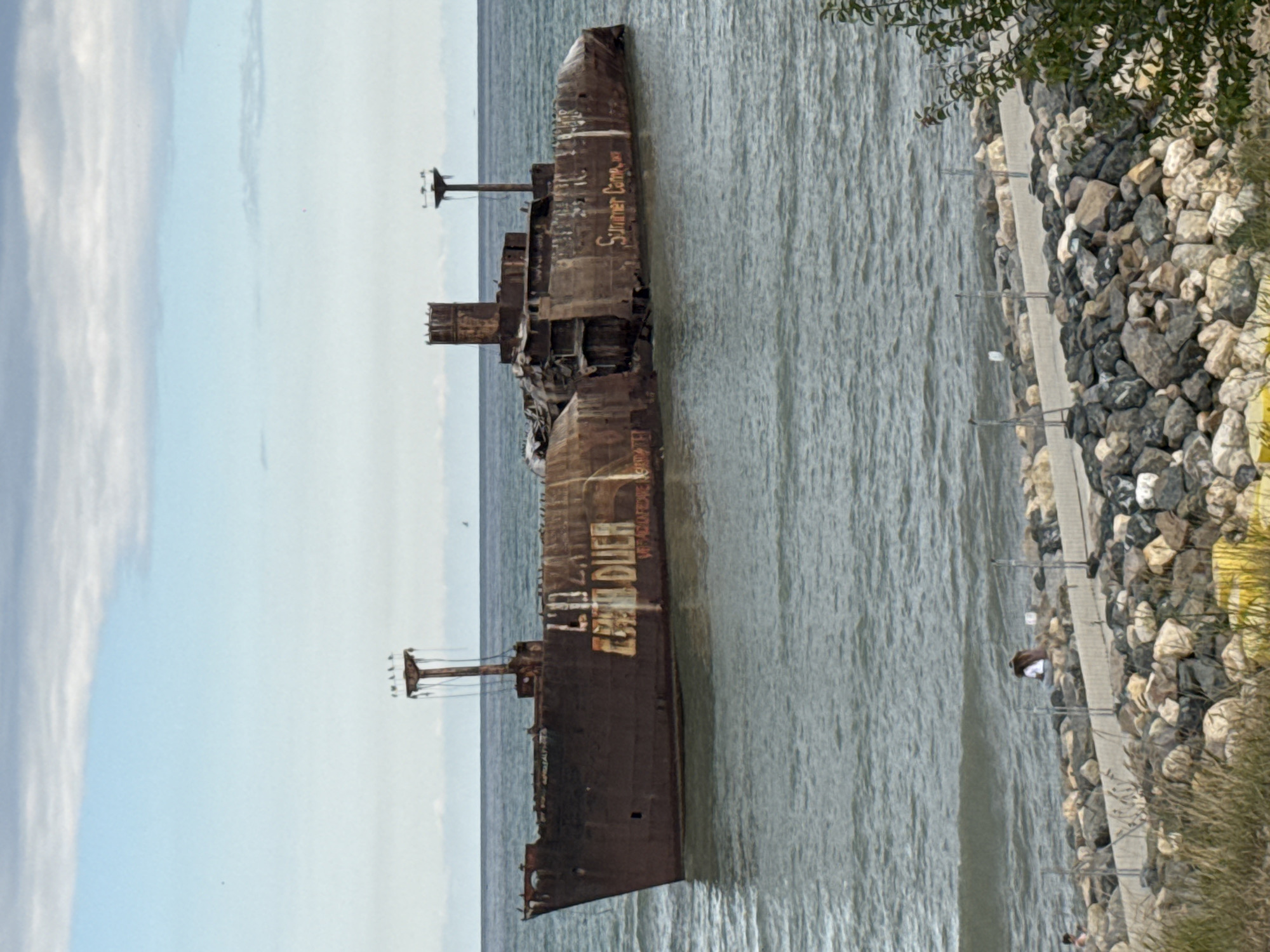 The image depicts the shipwreck of the Evangelia, a ship that ran aground off the coast of Costinești, Romania. The rusted hulk of the vessel is partially submerged in the sea, with visible damage and debris. The ship has graffiti on its side, including the words "EVANGHELIA" and "Summer Camp". In the foreground, a rocky shoreline with a paved walkway is visible, with a few people on it. The sea is choppy, and the sky is overcast. The overall scene is one of decay and abandonment, with the shipwreck serving as a stark reminder of the sea's power.