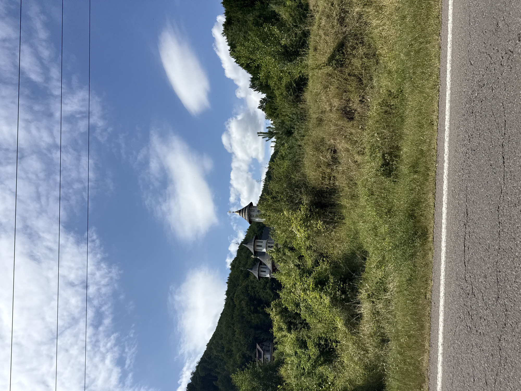 The image shows a view from a roadside in Ferestrău-Oituz, Romania. In the foreground, there is an asphalt road with a white line marking its edge. Behind the road, a grassy slope rises, covered with dry and green vegetation. Further up the slope, nestled among dense green trees and bushes, is a church complex. The church features multiple spires topped with crosses. The background consists of a densely forested hillside under a bright blue sky with scattered white clouds. Several power lines are visible stretching across the top of the frame. No people are visible in the image.