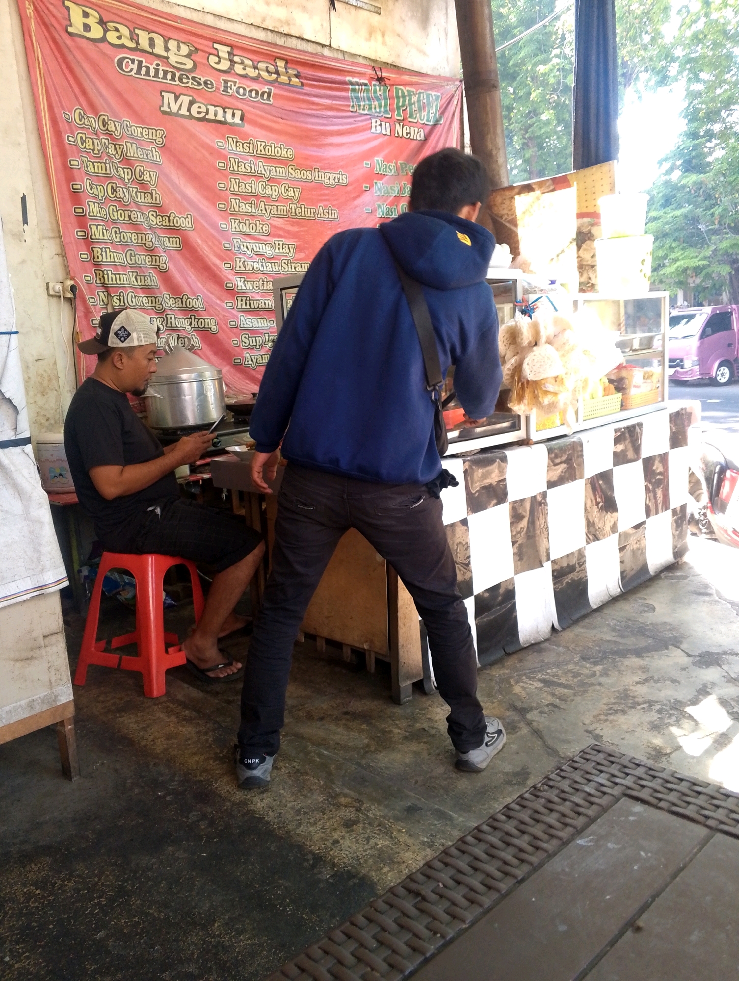 The image depicts an outdoor scene, possibly a food stall or eatery, with a man sitting on a red stool and another man standing. The standing man, wearing a blue hooded sweatshirt and dark pants, is facing away from the camera, looking towards a food stall. The seated man, wearing a black t-shirt, black shorts, and a cap, is looking down at a smartphone. Behind the standing man is a food stall with a checkered black and white tarp along its base. A pink vehicle is visible in the background, suggesting a public area. A large banner with Indonesian text, possibly advertising "Bang Jack Chinese Food Menu," is prominently displayed on the wall. The environment appears to be somewhat worn, with a concrete floor and a wooden structure to the left. The lighting suggests it is daytime, with sunlight filtering through trees.