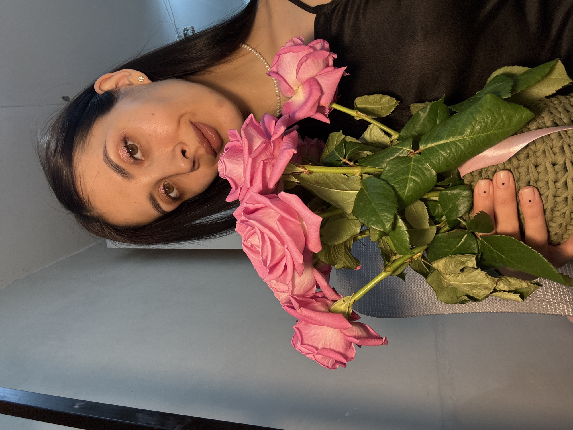 This image, captured in Votkinsk, Russia, features a young woman lying down and looking towards the camera, holding a bouquet of pink roses. She has long, dark hair and is wearing a dark top and a pearl necklace. Her makeup includes pink eyeshadow and glossy lips, and she has a small, light-colored stud earring in her right ear.
The bouquet of several vibrant pink roses, complete with their green leaves, is arranged across her chest and to her right. A light pink ribbon is tied around the rose stems. Her fingers, visible on the right, show light nail polish and small dark dots on two nails, resting on what appears to be a green, textured, knitted fabric or blanket.
The background is a plain, light-colored wall or surface, with a dark linear element visible in the bottom left corner. The lighting is soft, indicating an indoor setting. The woman exhibits a calm and gentle expression, with a subtle smile. There is no visible text in the image.