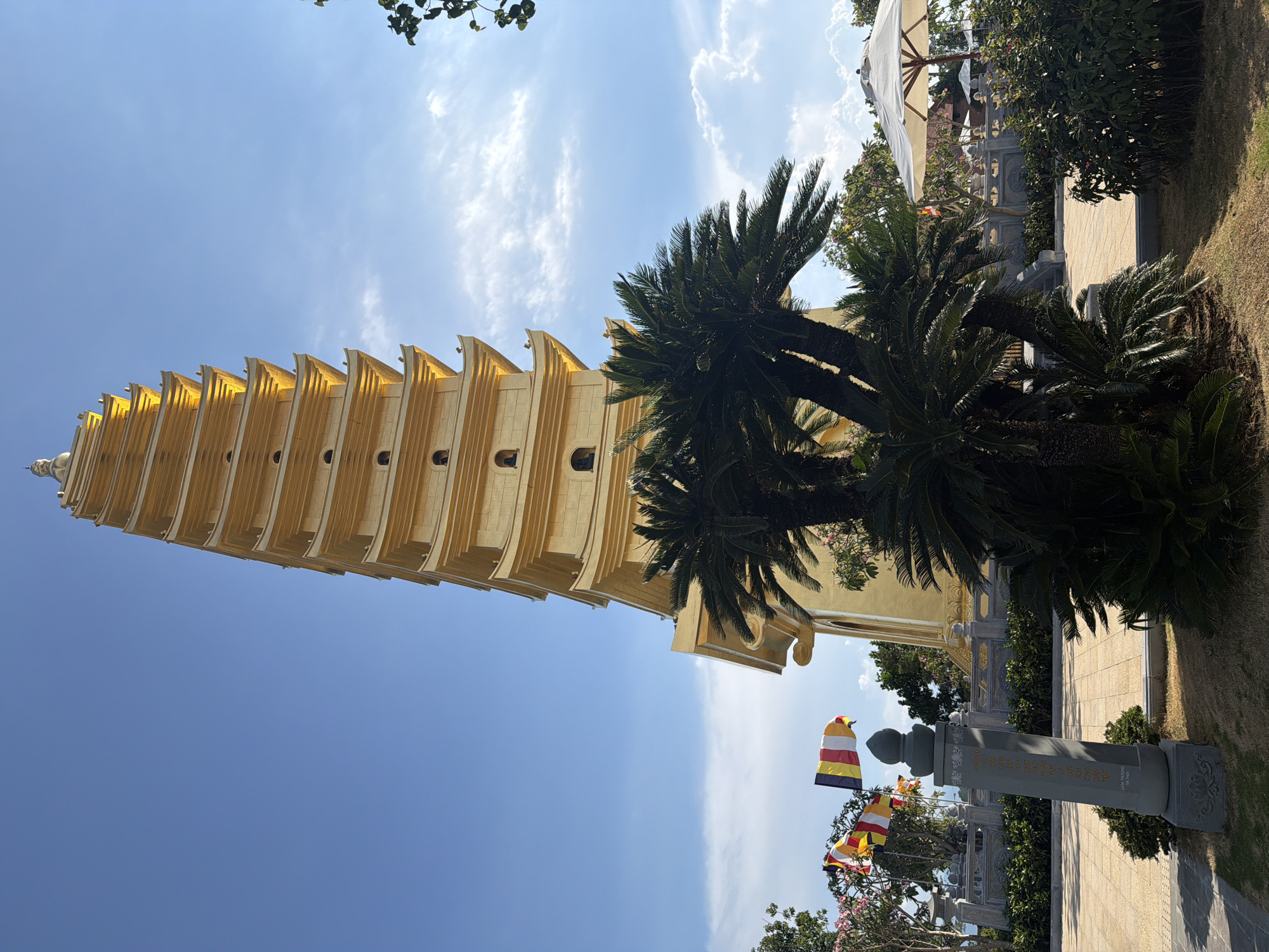 The image shows a tall, golden pagoda with multiple tiers, set against a blue sky with scattered clouds. The pagoda stands in a park-like setting, with palm trees and other vegetation visible. Buddhist flags are displayed near a stone pillar with inscriptions. The location is Nam Nha Trang Ward, Vietnam.