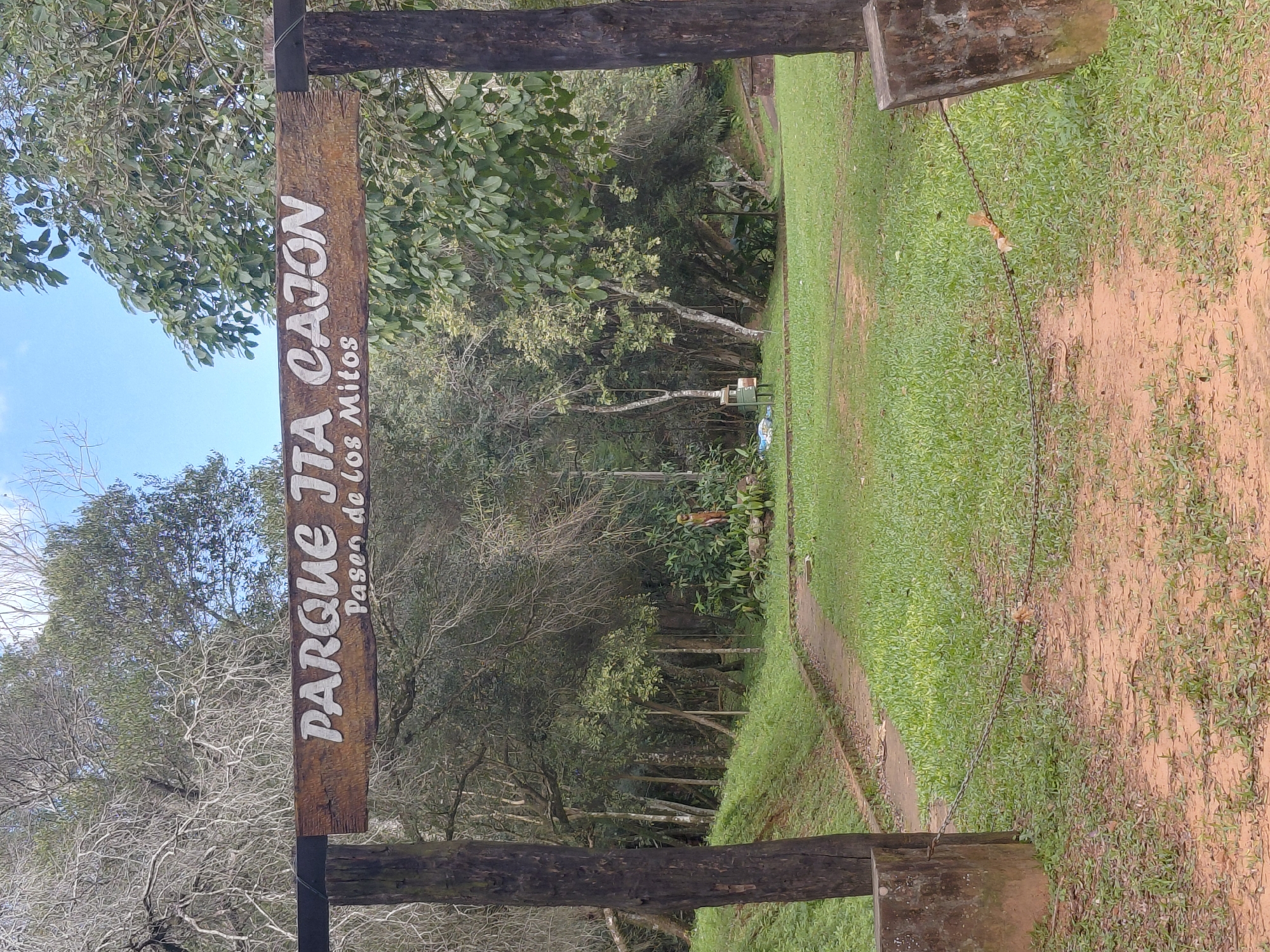 The image depicts the entrance to a park, as indicated by the sign that reads "PARQUE JTA CAJON" and "Paseo de los Mitos". The park appears to be located in a natural, wooded area with lush green grass and trees. A dirt path leads into the park, bordered by a chain fence in the foreground. The overall atmosphere is serene and inviting, suggesting a place for leisure and exploration. The park is located in the city of Trinidad, Paraguay.