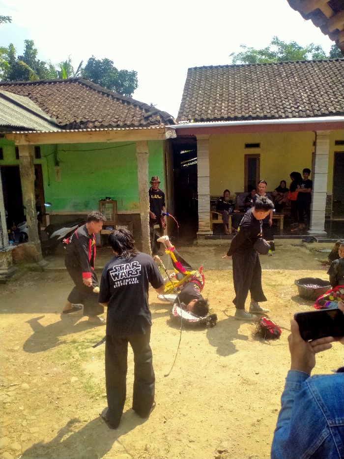The image depicts an outdoor performance or ritual taking place on a dry, light-colored dirt ground in Tawangsari, Indonesia. In the foreground and midground, four individuals are actively engaged. One person, wearing a black t-shirt with "STAR WARS THE EMPIRE STRIKES BACK" visible on the back, stands facing a second individual who is lying prone on the ground, face down, with their legs elevated and bent at the knees. This prone individual is adorned in colorful fabrics (red, yellow, green, blue). A thin rope extends from the standing "Star Wars" individual towards the prone figure, and a dark, possibly artificial hairpiece rests near the prone figure's head. To the left, a third individual in black attire with red accents strikes a dynamic pose, facing the prone figure. A fourth person, also in black, stands to the right of the prone figure, observing.
In the background, two single-story houses with brown tiled roofs are visible: one with light green walls to the left and another with light yellow walls to the right. Between these houses, a person in a dark shirt and a woven hat stands in a doorway, holding a bow, and observing the scene. On the porch of the yellow house, several individuals are seated and standing, acting as spectators. To the far right in the foreground, a person's hands and part of a blue denim jacket are visible as they hold a smartphone horizontally, seemingly recording the event. A black basin containing dark material and some red fabric rests on the ground near the performance area. The scene is brightly illuminated by direct sunlight, casting distinct shadows.