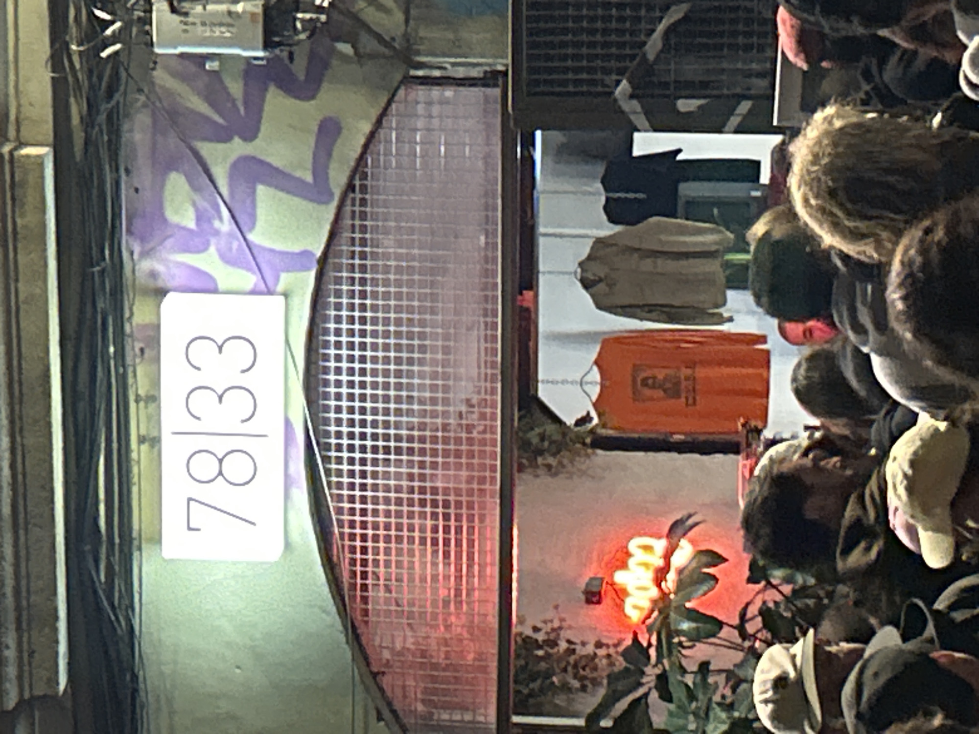 The image shows the exterior of a building in what appears to be a city environment. The address "78/33" is prominently displayed on a vertical, illuminated sign on the left side of the frame. The building facade next to the address is covered in light purple graffiti. A rounded, tiled window or doorway is next to the graffiti.
To the right of the window is a reflection of an interior space or scene. An orange sign or object is visible, along with a light-up sign and foliage. The reflection also shows several people grouped together.
The lack of specific architectural details and the generic urban elements do not provide enough evidence to definitively confirm that this is Barcelona, Spain. However, the style of the building and street scene could be suggestive of that location.