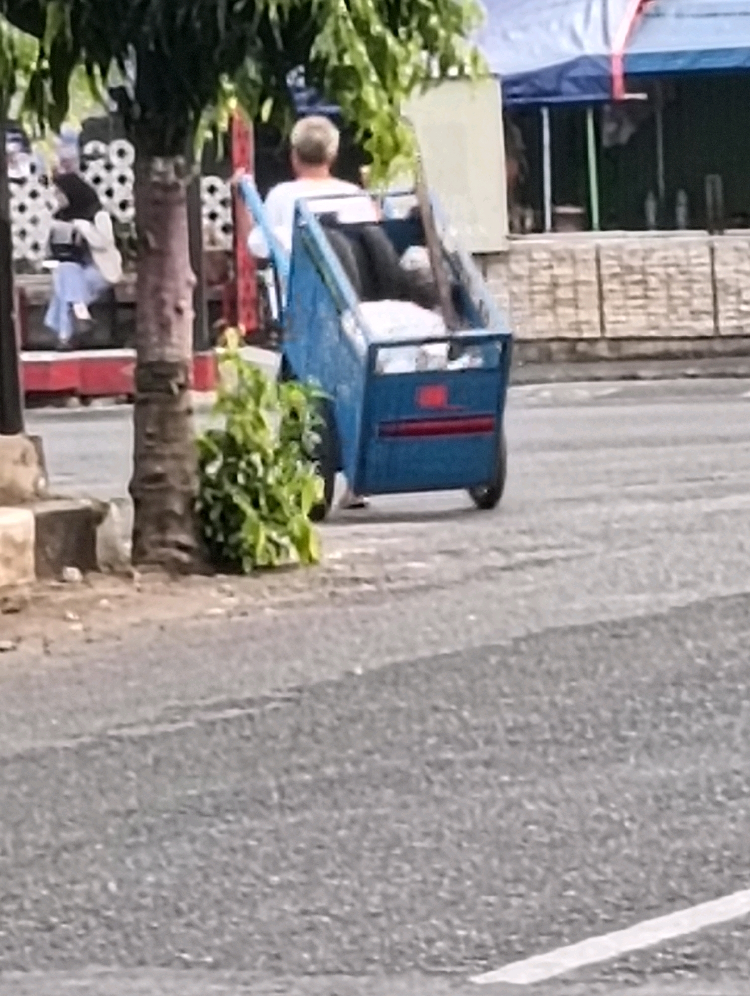 The image shows a blurry outdoor scene in Purworejo, Indonesia. In the foreground, a person with light-colored hair is operating a blue tricycle, which appears to be carrying a passenger or goods in the back. The tricycle has a red stripe on its cargo area. To the left of the tricycle, a tree trunk with rough bark is visible. Behind the tricycle, there is a building with a blue and white awning. In the far background, there are some indistinct figures and structures. The ground appears to be paved, with some of it looking like asphalt. The overall lighting suggests it is daytime.