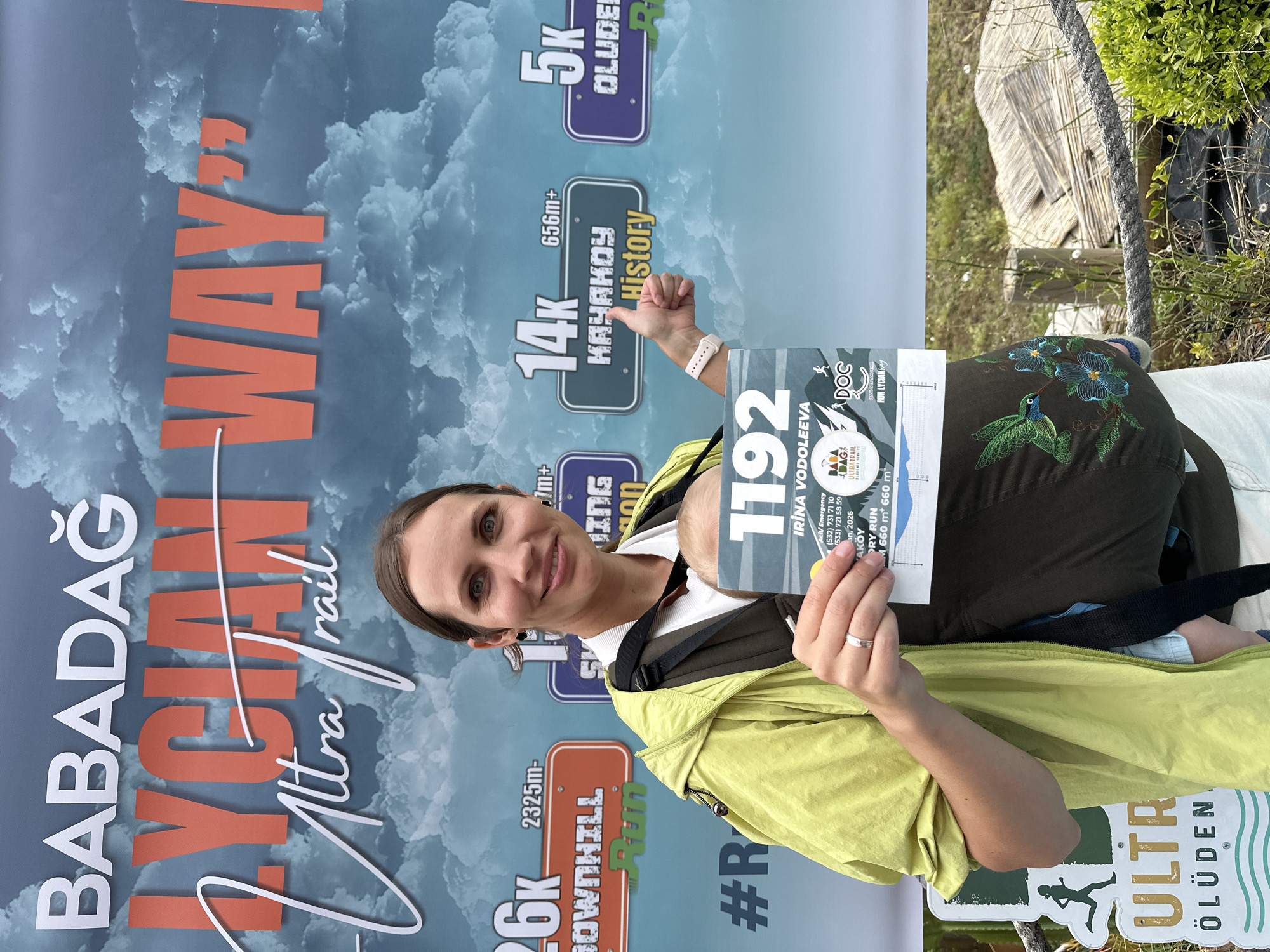 A smiling woman, wearing a bright yellow-green jacket over a white top and dark embroidered trousers, poses for a vertical portrait. She carries a baby in a front carrier, whose head is partially visible. The woman holds a race bib numbered "1192" with the name "IRINA VODOLEEVA" and details for the "BABADAG ULTRATRAIL RUN LYCIAN WAY," including emergency contacts and an elevation profile for an "AKÖY DRY RUN" (660m ascent, 660m descent).
The background is dominated by a large event banner, displaying "BABADAĞ LYCIAN WAY Ultra Trail" in prominent lettering against a cloudy sky. The banner also features various race distances and their characteristics: "26K DOWNHILL Run" (2325m- elevation), "14K KAYAKOY History" (656m+ elevation), and "5K OLUDEN". Visible beyond the banner are outdoor elements such as green foliage, a wooden structure, and a thick rope, suggesting a natural, possibly mountainous setting. The woman's happy expression and the event context indicate she is participating in or associated with a trail running event in Fethiye, Turkey, specifically around the Babadağ mountain and the Lycian Way trail. The scene is brightly lit, indicating daytime.