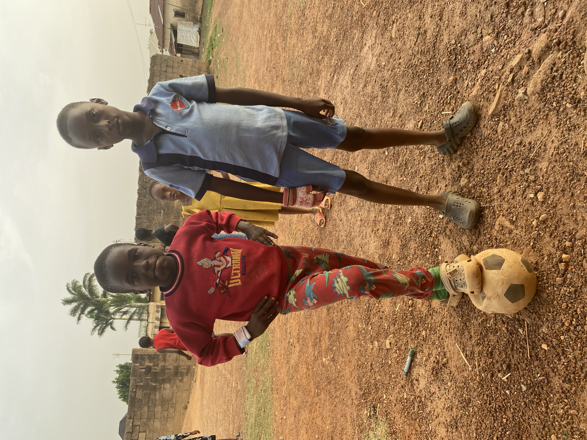 Two young boys are prominently featured in an outdoor, eye-level shot, likely in Bukuru, Nigeria. The scene is set on dry, reddish-brown dirt and gravel, indicative of an unpaved area. The sky above is overcast or hazy, diffusing the light evenly.
On the left, a boy with dark skin and short dark hair wears a red long-sleeved sweatshirt adorned with "ULTRAMAN ROBOT" text and an image of the Ultraman character. His red pants are patterned with what appear to be dinosaurs or similar creatures. He has a slight smile, his left hand on his hip, and his right foot rests on a worn, light-colored football (soccer ball) with dark hexagonal patches. He wears light-colored, open-toed shoes over socks.
Standing to his right, another boy with dark skin and short dark hair wears a light blue polo shirt with dark blue trim and a small emblem on the chest, paired with dark blue shorts. He has a neutral expression and wears dark grey Crocs-like shoes.
Behind these two, another child in a yellow top is partially visible. In the background, simple structures made of concrete blocks and some shanty-style buildings with corrugated iron roofs can be seen. A tall palm tree and other sparse vegetation are also present. On the ground near the football, a small green marker or crayon lies.
The boys appear to be posing for the camera, possibly during a casual playtime activity, given the presence of the football. The overall atmosphere suggests a relaxed moment in a community setting.