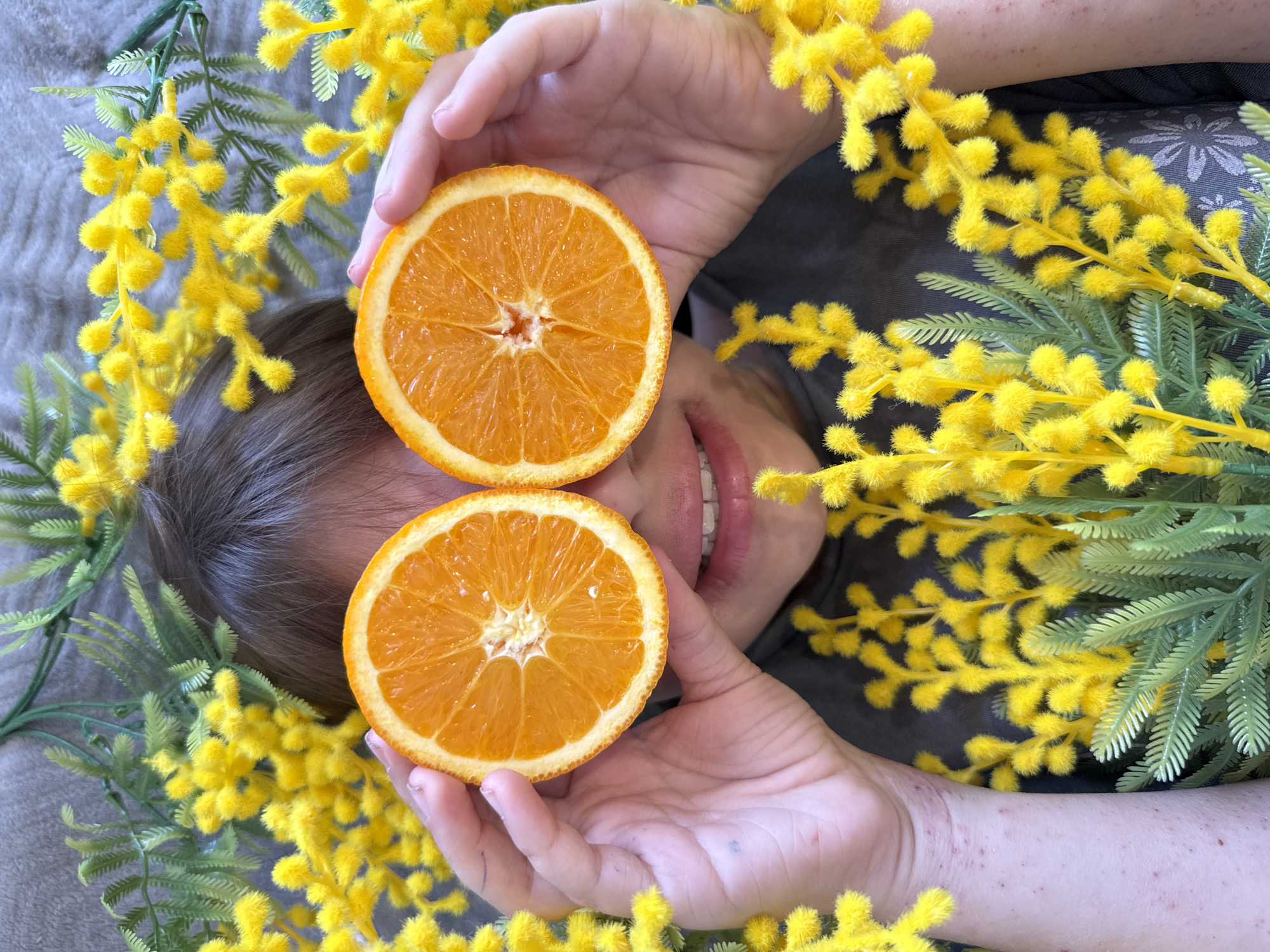 An overhead shot depicts a person lying down, playfully holding two halves of a cut orange over their eyes. The person's face is partially visible from the nose down, revealing a wide, joyful smile with white teeth. Their brown hair frames the upper part of their head.
The person's hands hold the bright orange fruit, which is sliced in half to show the juicy segments. They are surrounded by abundant artificial mimosa flowers, characterized by their vibrant yellow fluffy blooms and green fern-like leaves, creating a bright and cheerful frame around the person. The background appears to be a soft, textured gray fabric, possibly a blanket or carpet, suggesting an indoor setting.
Notable details include the person's fair skin, with some small red marks or spots visible on their arms. One hand shows a small dark dot. The overall mood is lighthearted and playful, emphasized by the person's smile and the vivid colors of the oranges and flowers. There is no visible text in the image.