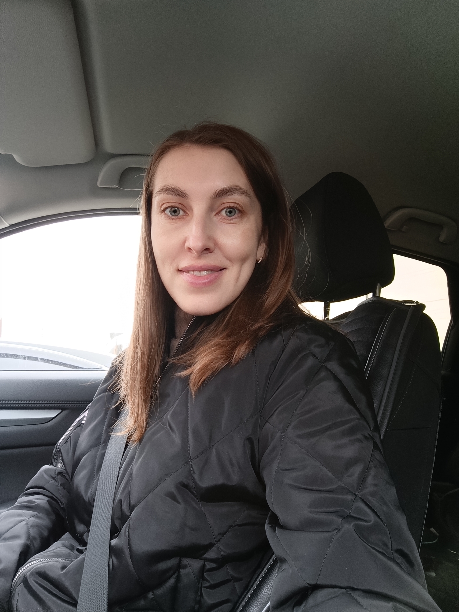 A woman is seated in what appears to be the driver's seat of a car, captured in a portrait-style shot. The setting is indoors, within a vehicle, with the provided context placing it in Chaikovsky, Russia.
The subject is a woman with light brown hair, blue eyes, and fair complexion, appearing to be in her 20s or 30s. She is looking directly at the camera with a subtle, friendly smile. She wears a black quilted jacket, which features a diamond pattern and zippers. A dark grey seatbelt is fastened across her chest, indicating she is either driving, a passenger, or preparing for travel. A small, silver earring is visible in her right ear. Underneath the jacket, a light brown or beige high-neck garment is partially visible.
The car's interior is largely dark-toned, with black or dark grey leather seats, a headrest visible behind the woman, and matching door panels. A grey sun visor and parts of the overhead console are visible in the upper portion of the frame. Through the driver's side window, the outside environment is bright and diffused, suggesting an overcast or cloudy daytime. No distinct landmarks or specific details of Chaikovsky are visible outside, only a bright, featureless sky and faint distant shapes. No text is visible in the image. The overall impression is a casual photograph taken inside a car on an ordinary day.