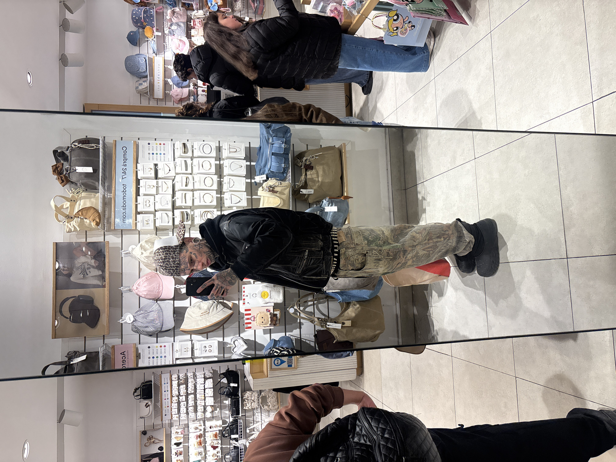 The image shows the reflection in a mirror of a retail store interior. Inside the store, various merchandise displays are visible, including shelves with hats, bags, and jewelry. Two women are seen browsing the merchandise. In the foreground, the reflection of a person with tattoos on their face and arms is taking a picture with their smartphone, as well as another person wearing a black quilted jacket. The store's clean, tiled floor and bright lighting suggest a modern and well-maintained retail environment. While the image provides details about the store's interior and merchandise, there is no information to confirm that the location is in the city of Buenos Aires, Argentina.