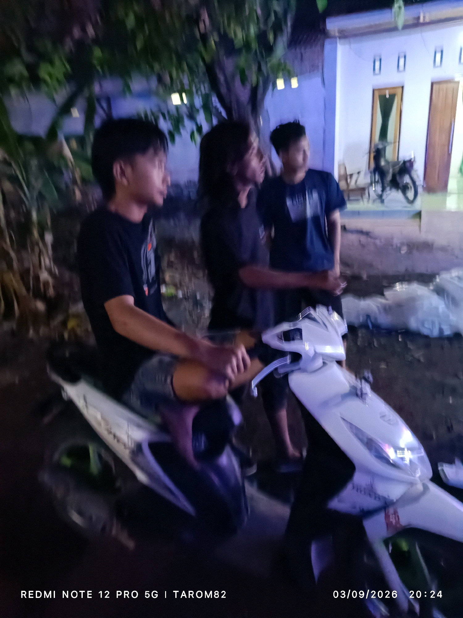 In Rejoagung, Indonesia, at night, three young males stand near a white scooter in front of a modest building. The individual on the left, seated on the scooter, wears a black t-shirt and shorts, looking away from the camera. The male in the center, standing, is dressed in a black t-shirt and dark shorts, facing left. The third male, standing to the right, wears a dark blue t-shirt and dark shorts, looking toward the building. The building appears to be a residence with a light-colored facade, two doors, and three windows. A motorcycle is parked near the building's entrance. The ground is paved and appears wet, reflecting some of the ambient light. The overall scene is dimly lit, with some artificial lighting contributing to the nocturnal atmosphere. A timestamp at the bottom of the image reads "03/09/2026 20:24."