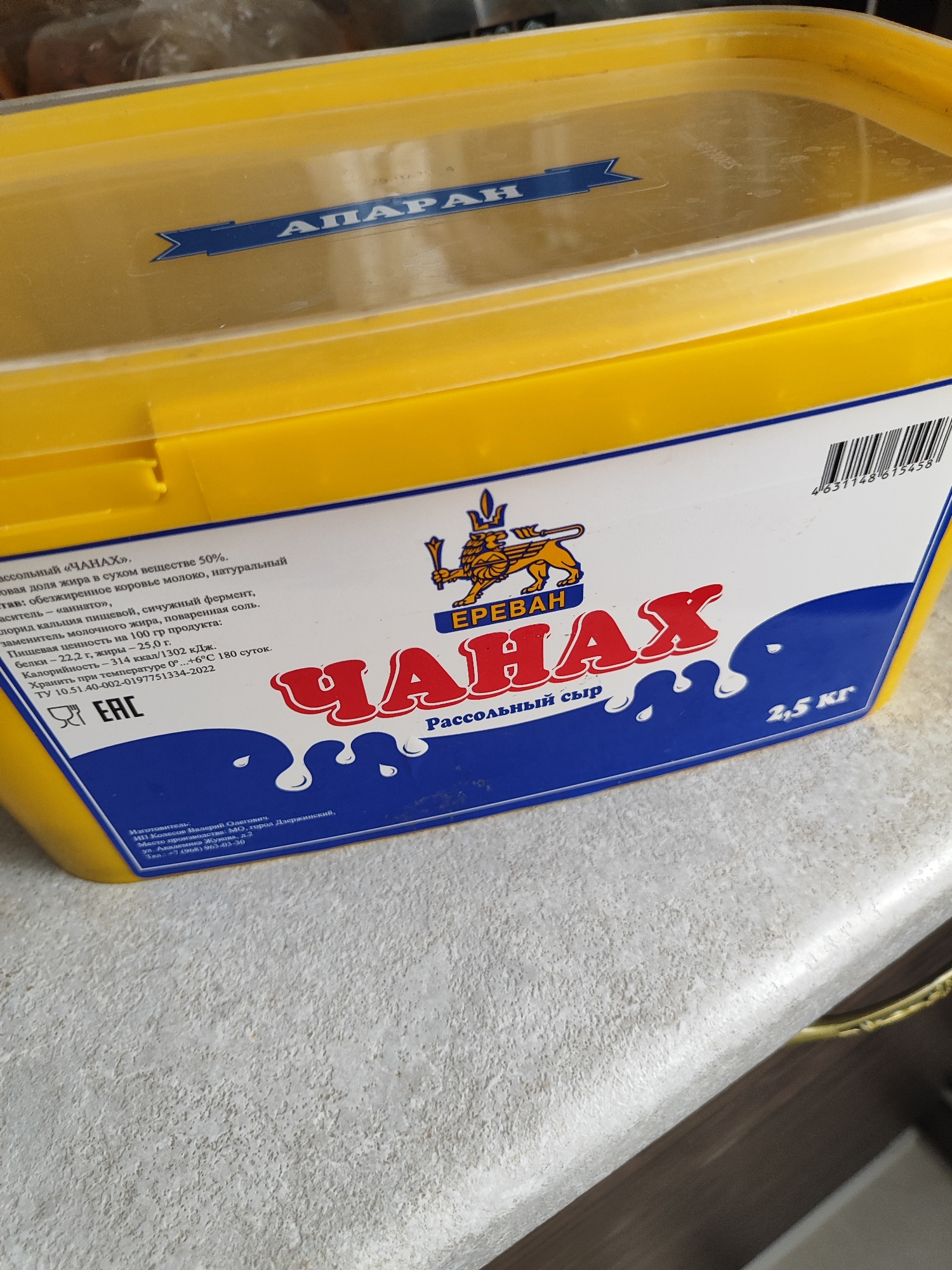 A bright yellow rectangular plastic container with a transparent lid is positioned on a light gray countertop. The lid has a blue emblem with white text that reads "АПАРАН". The front of the container features a blue label with white lettering and a graphic of a lion-like creature. Prominently displayed on the label is the word "ЧАНАХ" in large red letters, with "Рассольный сыр" in smaller white text below it. To the right, the text "2,5 кг" is visible in white. On the left side of the label, there is text detailing the product's ingredients, nutritional information, and manufacturing standards, along with an "EAC" logo. Below this, there is smaller text indicating the manufacturer's details and contact information. A barcode is visible on the upper right side of the label. The overall lighting suggests the image was taken indoors.