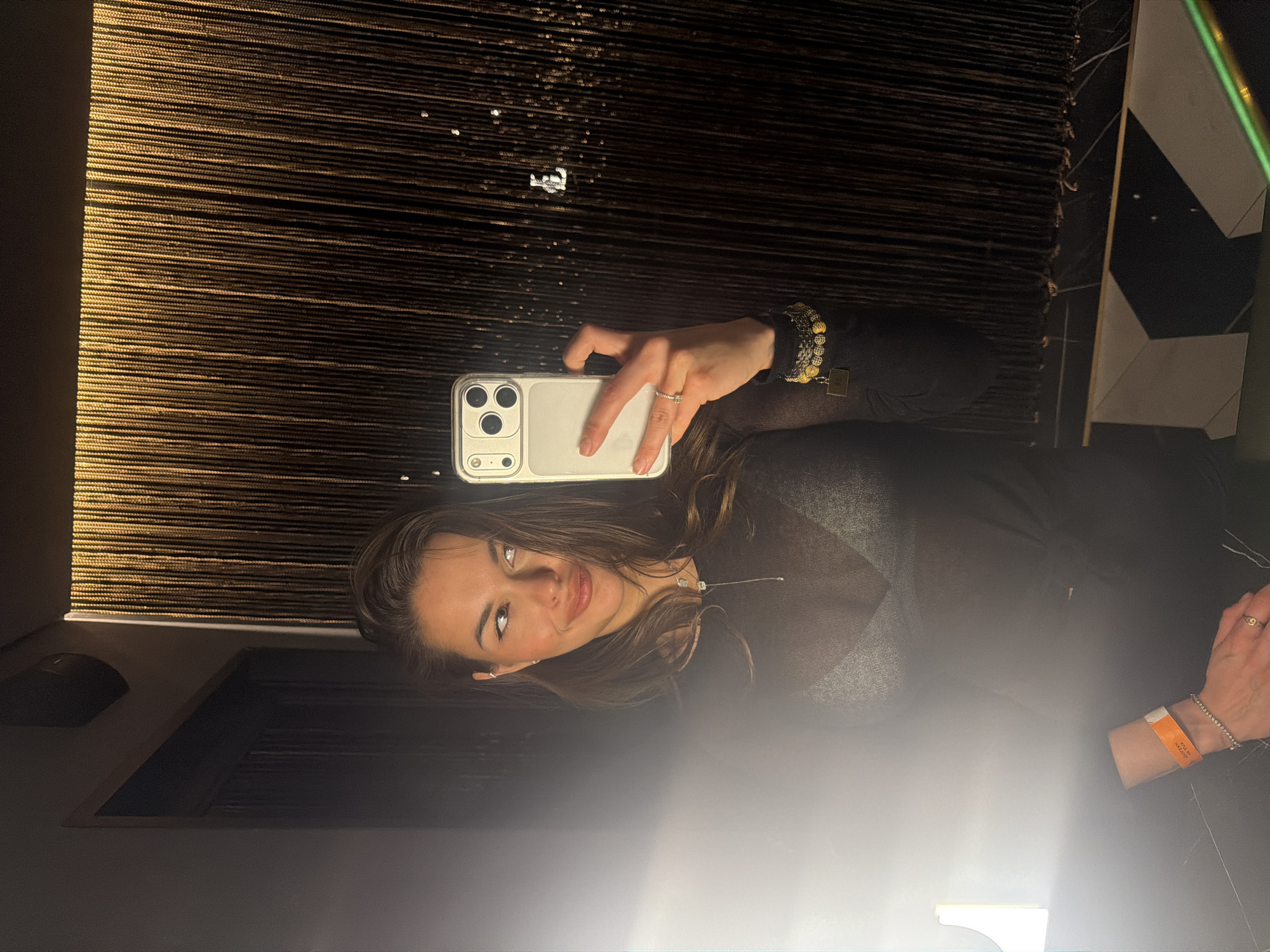 A woman is taking a selfie in what appears to be a restroom or a stylish interior space. She's holding an iPhone, which captures her image with the background in focus. The background has a textured, curtain-like design and a geometric pattern. An orange wristband and jewelry are visible on her wrist. The lighting is dim, with a bright reflection suggesting a light source is pointing towards her. There are no recognizable landmarks to determine the specific location in Krasnoyarsk, Russia.