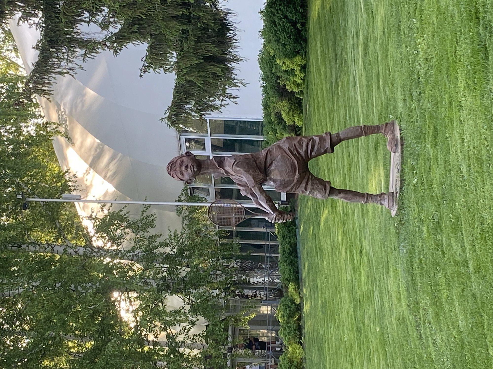 A bronze statue of a young boy playing tennis stands on a lawn in front of a building in Izhevsk, Russia. The building has white walls and glass windows and doors. Trees and bushes surround the statue and building. The statue is a life-size representation of a boy in shorts and a shirt, holding a tennis racket. The boy is in a ready position, preparing to hit the tennis ball. The lawn is green and well-manicured. The statue is situated in a park or garden setting, and a few people are walking about in the background.