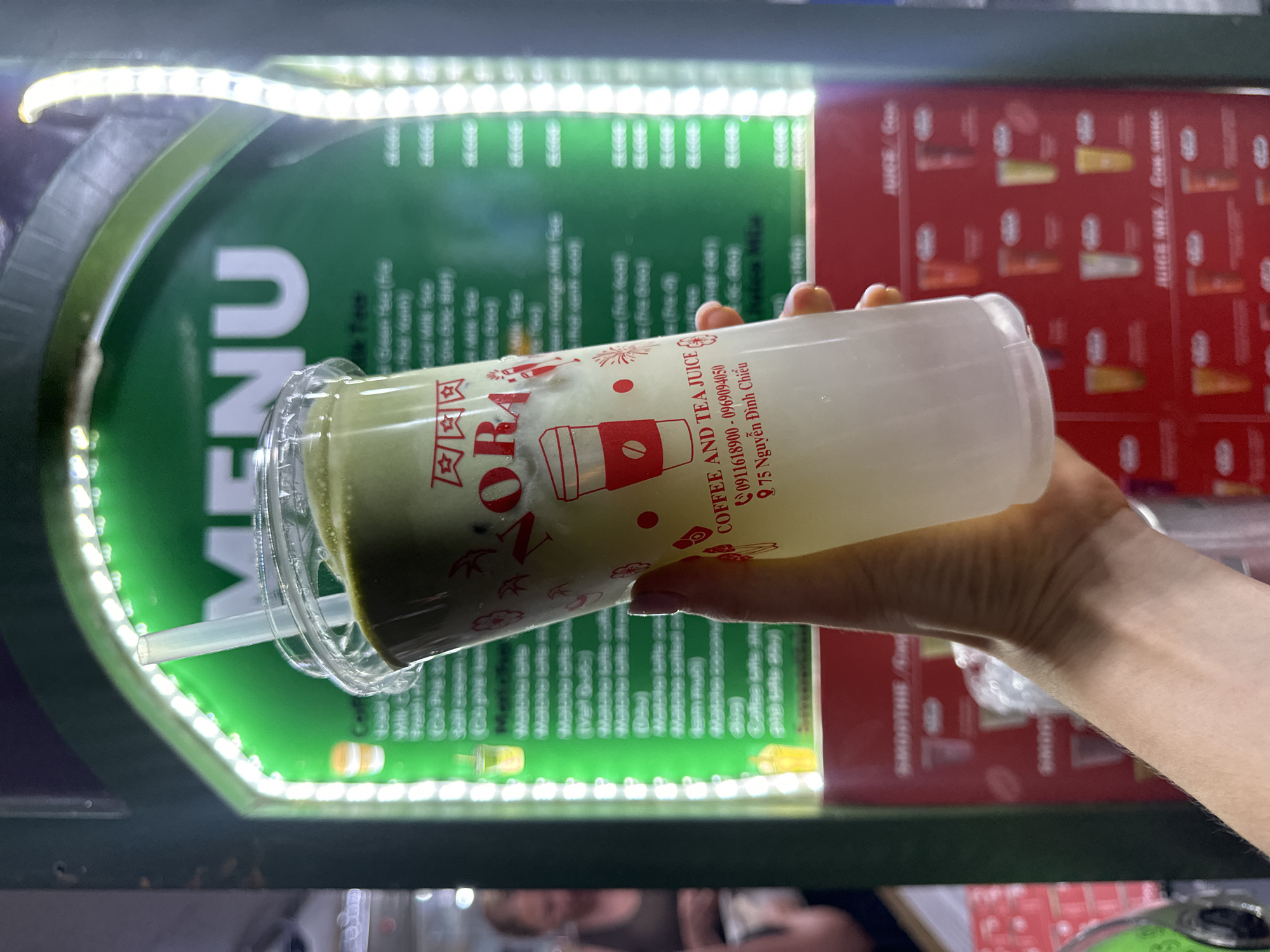 The image shows a person holding a layered drink in a clear plastic cup, set against a brightly lit menu board. The scene appears to be an outdoor food stall or small cafe at night, likely in Mũi Né, Vietnam, given the address and overall ambiance.
The main subject is a hand holding a plastic cup containing a beverage with distinct layers: a dark green layer at the bottom, possibly matcha or a green tea blend, and a thick white, frothy layer on top, likely milk or cream. A clear plastic straw is inserted into the drink. The cup is branded with red text that reads "ZORAA" (or "ZORA") and "COFFEE AND TEA JUICE." Below this, contact information includes phone numbers "0911618900 - 0969094050" and an address "975 Nguyễn Đình Chiểu," a street name in Vietnam.
In the background, a menu board is visible, outlined by bright white LED strip lights, suggesting it is nighttime. The menu board is divided into sections; the left side is green with the word "MENU" clearly visible, along with blurred text that includes "Tee" and "Green Tea." The right side of the board is red and features categories such as "JUICE / Cofe" and "JUICE MIX." The lighting creates a vibrant, urban street food atmosphere.