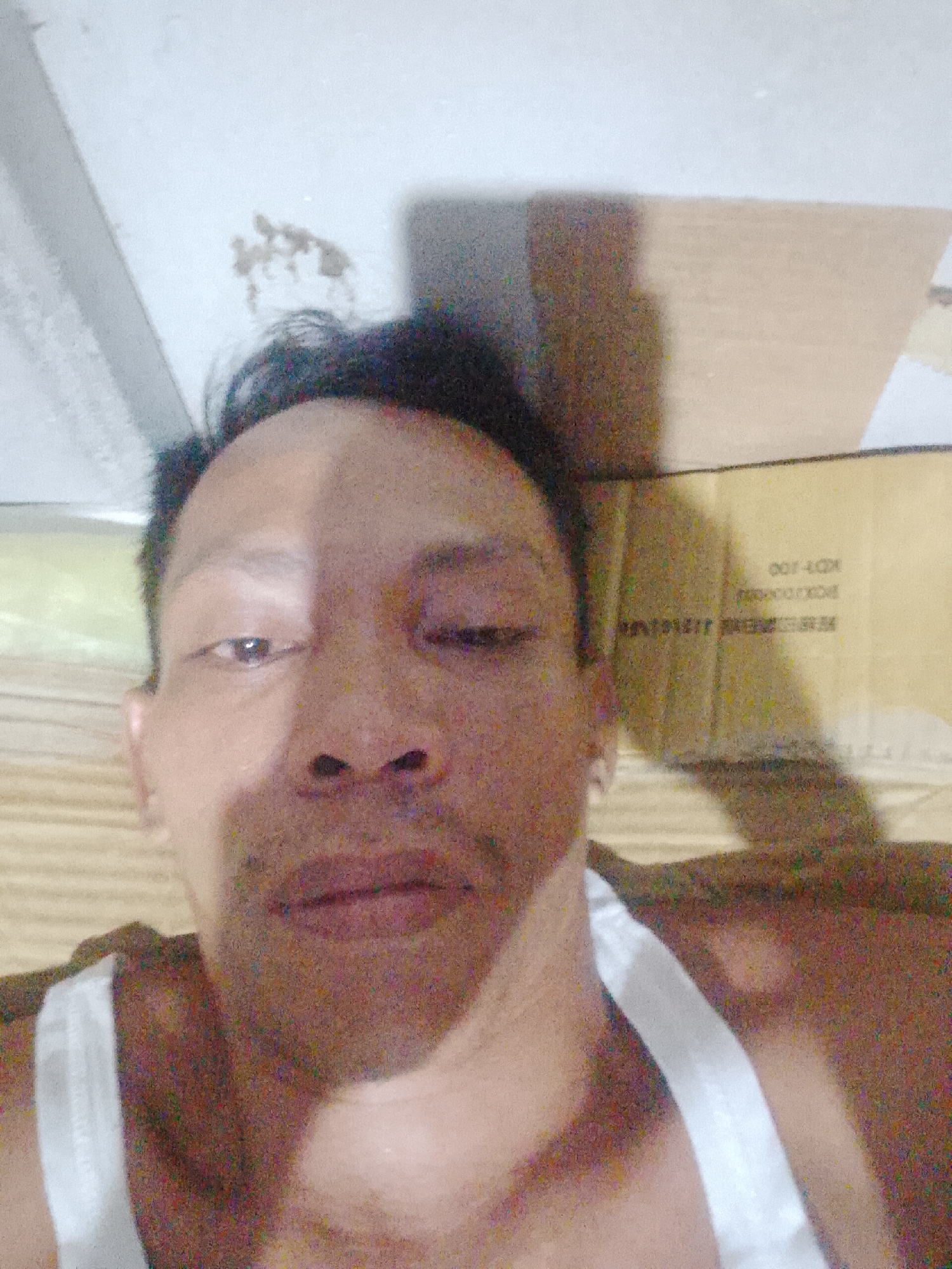 A close-up, low-angle selfie photograph depicts an adult male with dark hair and brown skin wearing a white tank top. The background includes a ceiling with some discoloration, a cardboard box with text that includes "KD7-100" and other illegible markings, and what appears to be a mattress with horizontal lines. The photograph's location is Bukit Ketok, Indonesia, but there is no further contextual information to establish the setting within the city. The subject's eyes appear to be partially closed, but this may be from the image quality. There is no indication of any other people, interactions, or activities occurring.