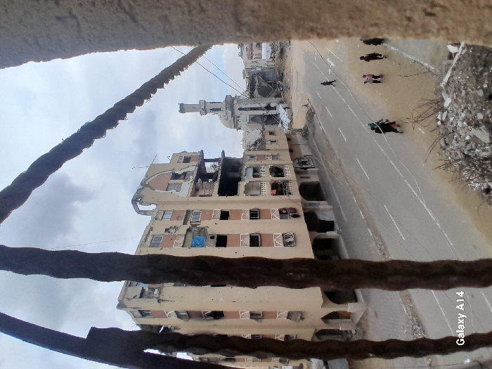 The image depicts a devastated urban landscape in Khan Yunis, Palestinian Territory, viewed from an elevated perspective through a frame of bent rebar and rubble. A multi-story building, heavily damaged with significant structural compromise, missing walls, exposed interiors, and broken windows, stands prominently on the left. Some window openings are covered with blue tarps. In the background, to the right of the damaged building, a mosque with a tall minaret and a dome is visible, also appearing to have sustained damage.
A paved road with white lane markings runs horizontally across the midground. Several individuals are present on or beside the road. One person walks alone on the left side of the street, while a group of approximately five individuals, including adults and possibly children, walks on the right side. One person in this group is dressed in dark clothing, including what appears to be an abaya. The sky is overcast. Debris and rubble are present in the immediate foreground, consistent with the viewpoint being from a damaged structure. A "Galaxy A14" watermark is visible in the lower-left corner.