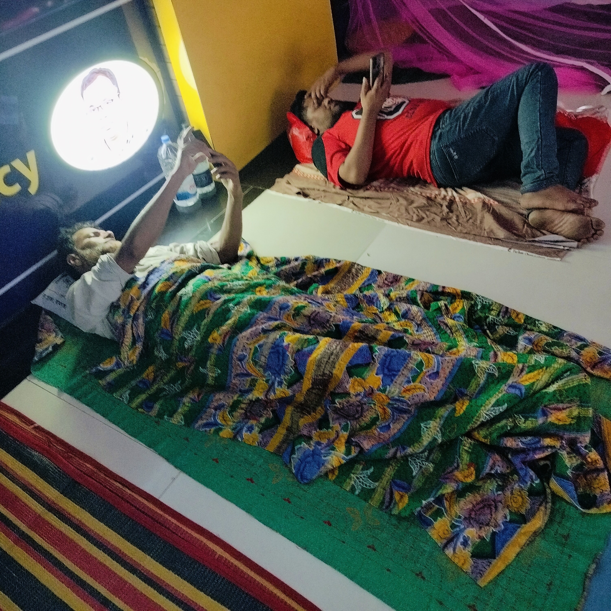 Two men are lying down in what appears to be a room in Dhaka, Bangladesh.
One man is lying under a patterned green, blue, and yellow blanket. He is using a cell phone. Next to him are two water bottles.
The second man is lying on a red cushion and brown mat. He is wearing a red t-shirt and blue jeans. He is also using a cell phone.
There is a framed picture on the wall, and a colorful net hanging above. The floor and wall surfaces are a light color. There is a striped mat on the floor.