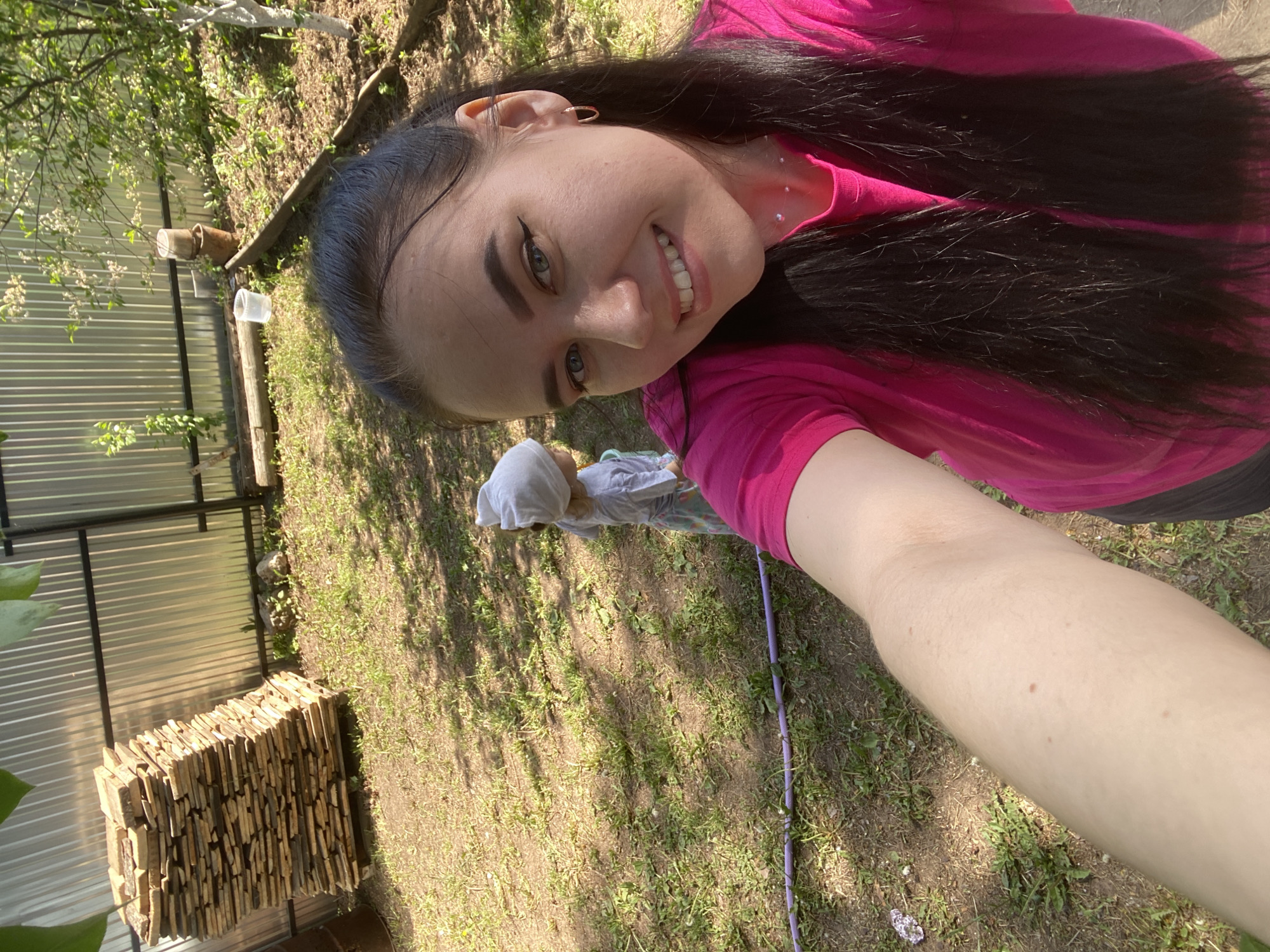 A selfie-style photograph shows a woman smiling at the camera. She has long, dark hair, fair skin, and wears a bright pink top. In the background, a small child wearing a light-colored hat is visible. The setting appears to be an outdoor area with grass, a stack of flat stones next to the fence, and what looks like a galvanized metal fence. The location might be in a backyard or garden area. The photograph was taken in Chaikovsky, Russia.
