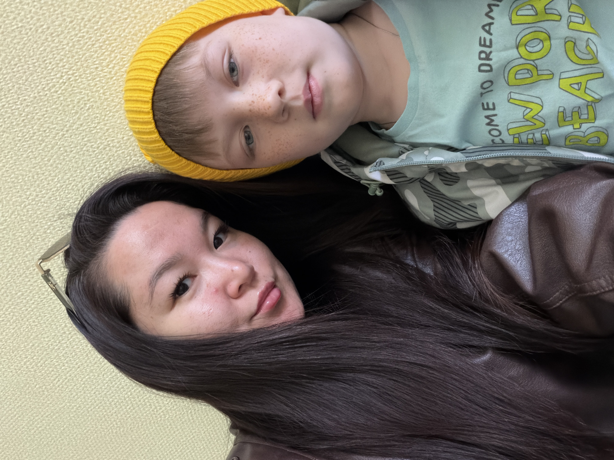A close-up shot depicts two individuals, a woman and a child, in an indeterminate indoor location, possibly in Unknown, Unknown. The woman, positioned lower in the frame, has dark, straight hair and appears to be wearing a brown leather jacket. A pair of glasses rests on her head. The child, positioned above and to the right of the woman, wears a yellow beanie and a light green shirt. The child has fair skin, freckles, and blue eyes. The backdrop is a textured yellow surface.
