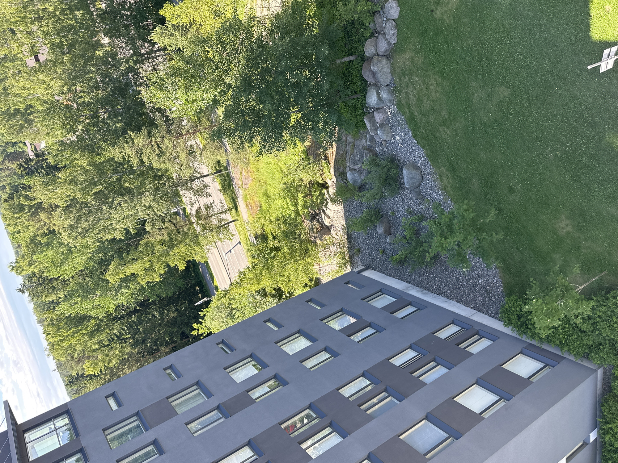 The image shows a modern, gray, multi-story building with numerous windows. The building is angled, and its facade faces the left side of the frame. The building has a dark gray roofline with a slanted overhang. Adjacent to the building, on the right side of the image, is a well-maintained green lawn. Behind the lawn, a landscape feature with large rocks and gravel is visible. Further in the background, a dense forest with lush green trees dominates the scene. A paved road with white markings can be seen winding through the trees. The sky above is partly cloudy with a bright, clear blue hue. The overall setting appears to be a residential area or a place with a blend of urban architecture and natural greenery. There are no people visible in the image.
