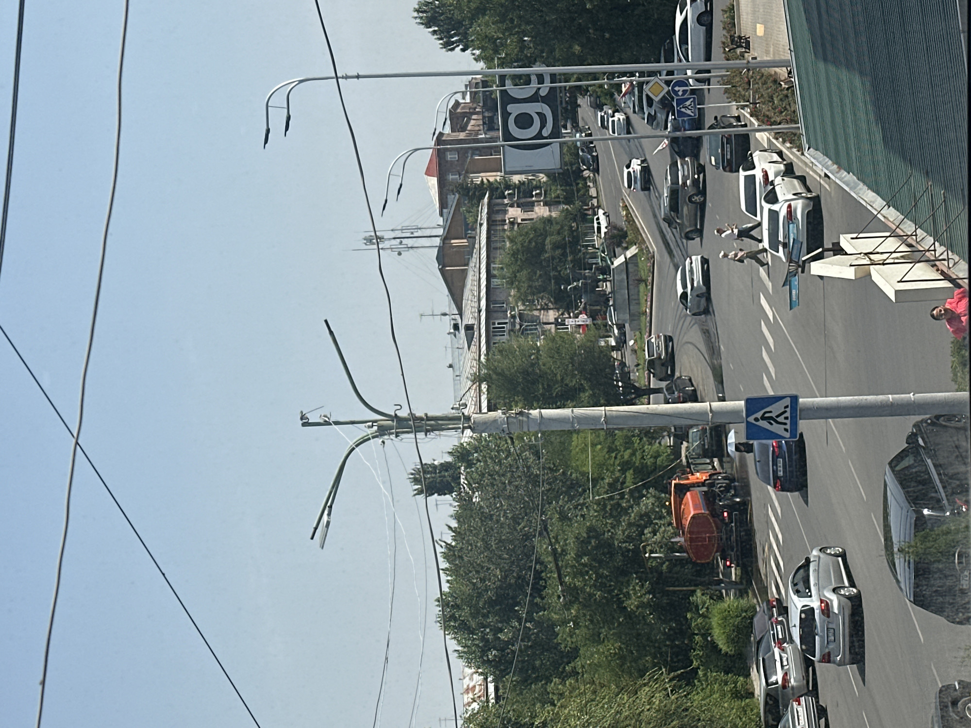 The image depicts a street scene in Gyumri, Armenia. A busy intersection is shown with numerous vehicles, including cars, SUVs, and a water truck. Pedestrians are crossing the street at a marked crosswalk, and a pedestrian crossing sign is visible. Buildings line the street, with trees and overhead wires interspersed throughout the urban landscape. The sky is clear and blue.