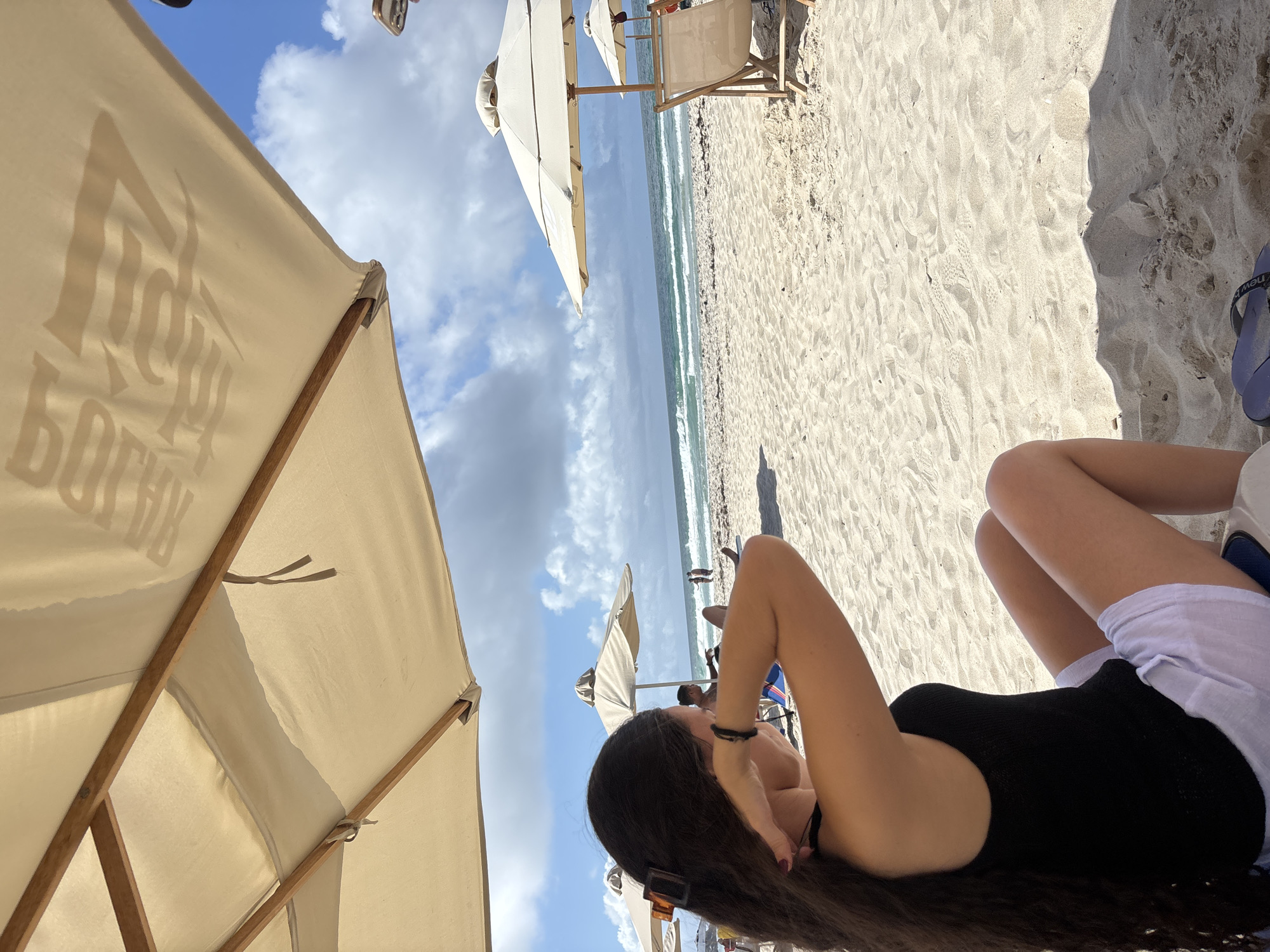 The image depicts a sunny day at a beach with light-colored sand. In the foreground, a person with long dark hair is seated on the sand, wearing a black tank top and white shorts, and appears to be adjusting their hair. The person is positioned under a large beige umbrella with wooden supports, which partially obscures the sky.
In the mid-ground, the beach stretches towards the ocean. Several other beach umbrellas and lounge chairs are visible, some occupied by people. The ocean has waves and appears to be a turquoise color under a blue sky dotted with white clouds. The scene conveys a relaxed atmosphere, typical of a beach resort.