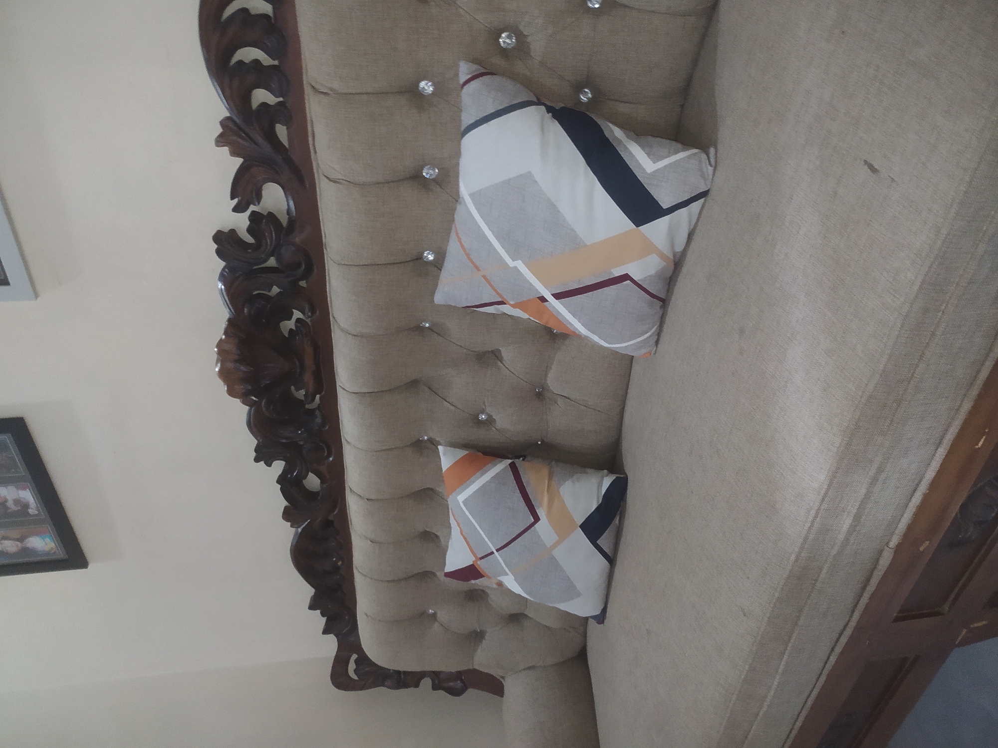 This is a medium-angle, eye-level shot of a tufted sofa with a dark wooden ornate headboard. The sofa is upholstered in a light beige fabric, and two square throw pillows with a geometric pattern in shades of gray, white, orange, and dark blue are placed on the seat. The background is a plain, light-colored wall. Above the sofa, a framed picture containing multiple people is visible. The image appears to be taken indoors, possibly in a living room. There are no people present in the image itself, and no actions are being depicted.