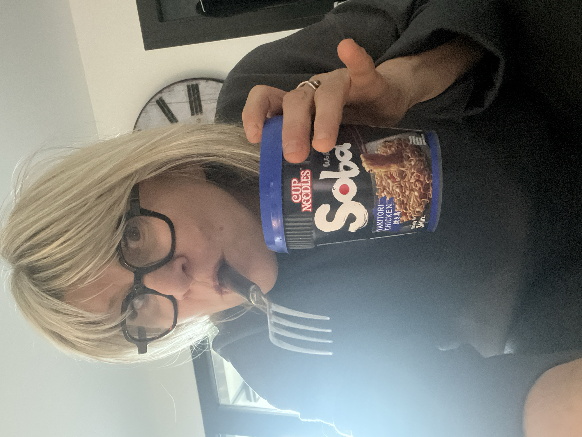 A woman with blond hair and glasses is holding a cup of Cup Noodles Soba Yakitori Chicken and eating noodles with a fork. A clock is visible in the background.