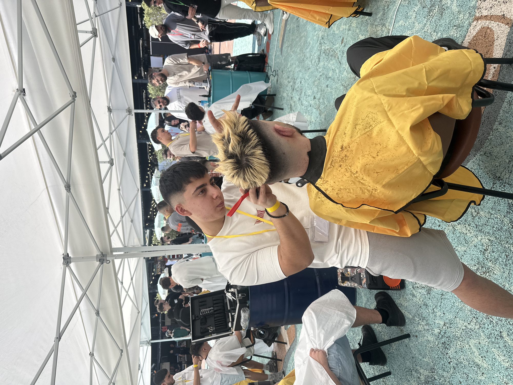 The image displays an outdoor barbering event taking place in Sofia, Bulgaria, characterized by multiple active stations under white canopy tents. In the foreground, a young male barber, wearing a white short-sleeved t-shirt, light grey shorts, a yellow wristband, and a clear identification badge, is seen from the chest up, focused on cutting a client's hair. He holds a red-handled straight razor in his right hand, while his left hand is positioned on the client's hair. The client, seated with his back to the camera, has distinctive blonde, spiky hair on top with sharply faded dark sides, and is draped in a yellow barber's cape visibly covered with hair clippings. They are positioned on a textured, light blue ground.
In the midground to the left, another barber attends to a client covered in a white cape. Beside this station, a dark blue metal barrel serves as a stand for an open black barber's case, revealing various tools including scissors, combs, and clippers. Further into the midground, several other men are visible, some standing and observing, others seemingly engaged in their own barbering tasks. The background shows more white canopy tents, additional people, and hints of an urban outdoor setting with darker structures and string lights. The overall atmosphere appears busy and professional, characteristic of a public skill demonstration or competition.