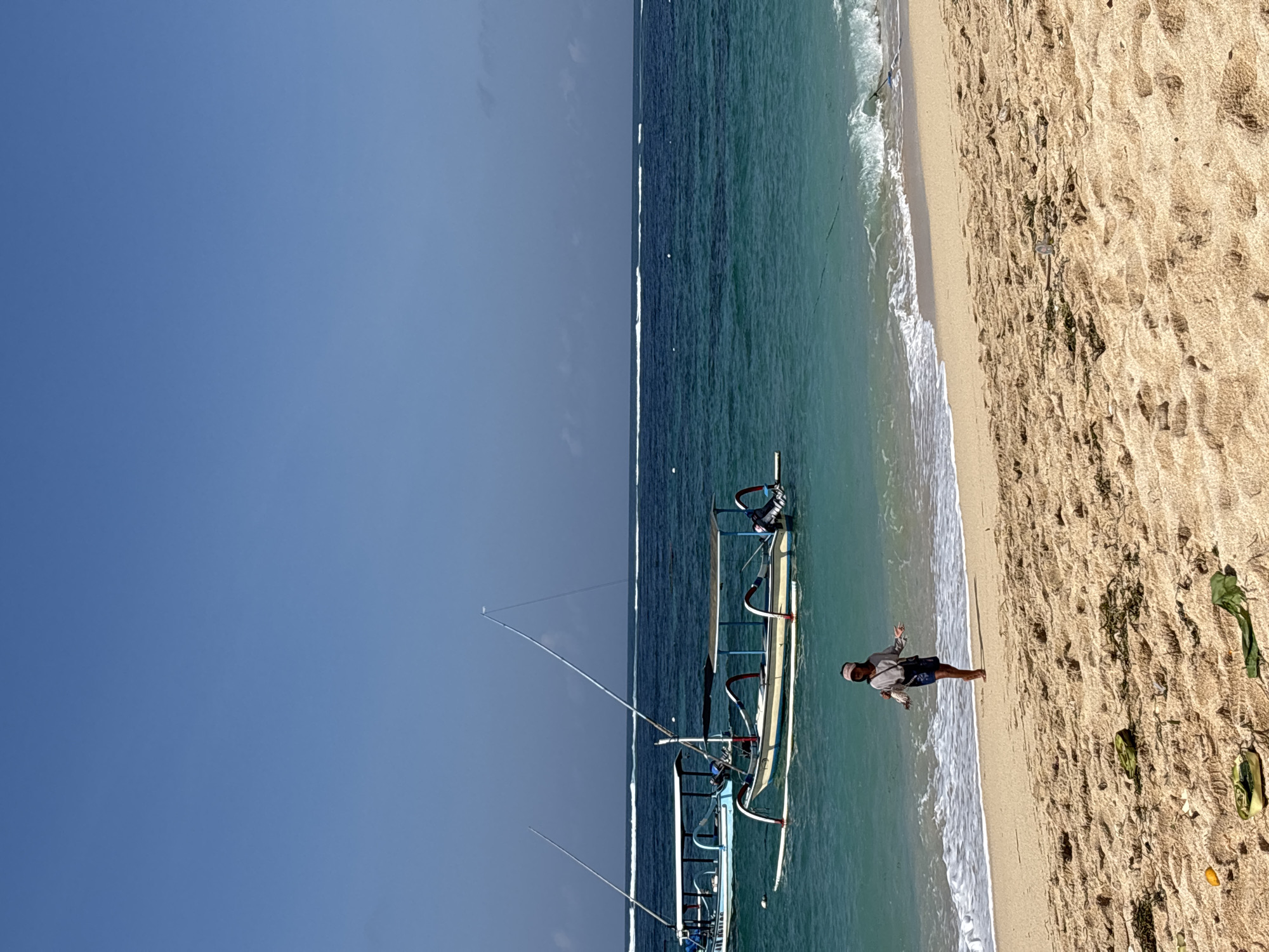 In Kuta Selatan, Indonesia, a serene beach scene unfolds under a clear blue sky. The sandy shore, dotted with scattered seaweed and a few discarded items, leads to the turquoise waters of the ocean. Two traditional Balinese jukung boats, characterized by their outriggers and tall masts, are anchored offshore. One boat, predominantly white with blue trim, is positioned closer to the shore, while another, a lighter cream color with red and blue accents, floats further out. A person, dressed in a light-colored long-sleeved shirt, dark shorts, and a pink head covering, stands near the water's edge, facing away from the viewer, seemingly walking along the beach. Gentle waves lap at the shore, creating a peaceful atmosphere. The horizon is marked by a thin line of white surf breaking in the distance.