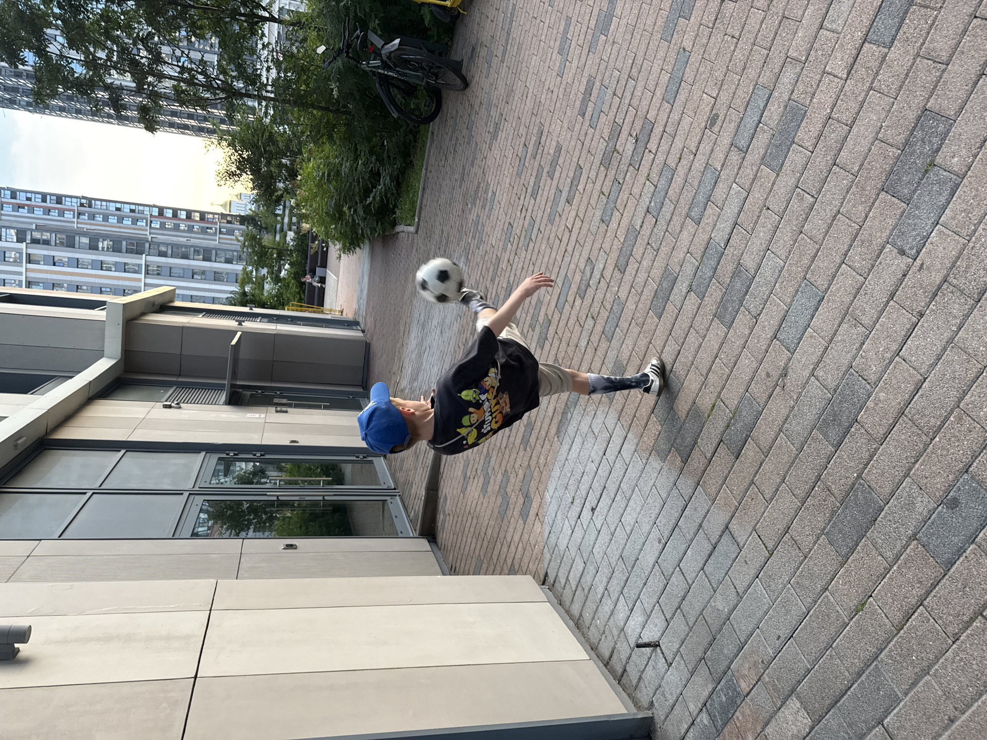 In Moscow, Russia, a young boy wearing a blue baseball cap and a t-shirt with cartoon characters is captured mid-action, kicking a soccer ball. He is dressed in light-colored shorts and dark socks with white athletic shoes. The scene unfolds on a paved courtyard, illuminated by natural daylight. Behind the boy, modern residential buildings with numerous windows dominate the background. Green bushes and trees are interspersed between the structures, and a bicycle is parked to the right of the frame. The architecture suggests a contemporary urban setting.