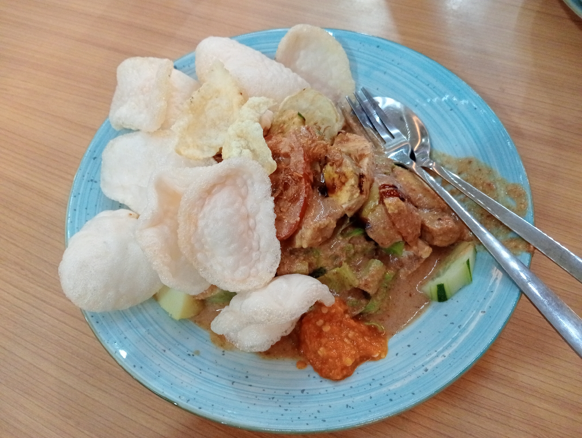 An overhead shot shows a blue speckled plate of Indonesian food on a wooden table. The food appears to be Gado-Gado, a salad of blanched vegetables, hard-boiled eggs, fried tofu and tempeh, and lontong rice cake, served with a peanut sauce dressing. A generous serving of krupuk (crackers) is on top of the salad. A fork and spoon are resting on the edge of the plate. The lighting is natural, suggesting the photo was taken indoors during the day. The setting is a casual eatery in Surabaya, Indonesia.