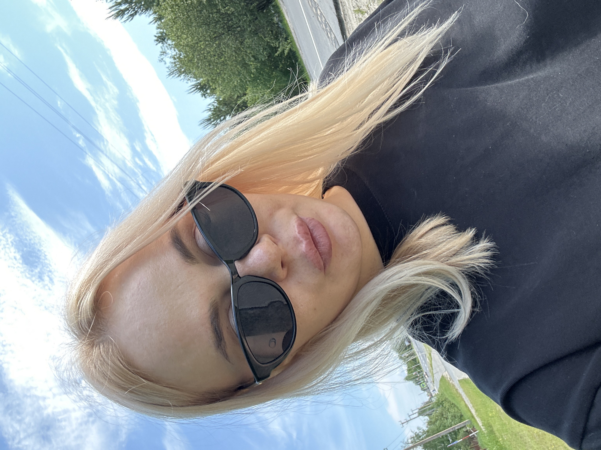 A woman with blonde hair and sunglasses is taking a selfie outdoors. She is wearing a black t-shirt. The background shows a blue sky with some clouds, trees, and a road.