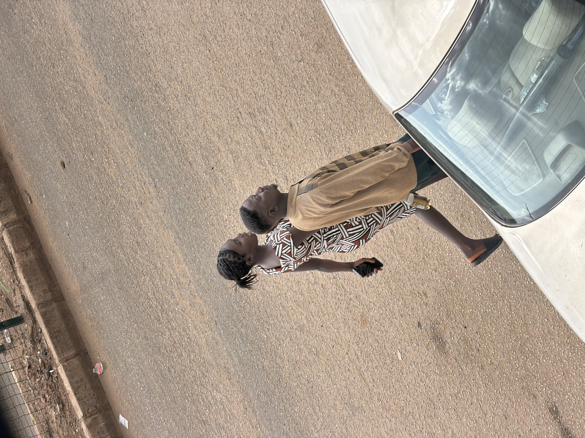 In Bukuru, Nigeria, two individuals, a woman and a man, walk along a paved road. The woman, on the left, wears a patterned dress and carries a dark object in her right hand. She has her hair braided into a ponytail. The man, to her right, is dressed in a light brown long-sleeved shirt with plaid accents on the shoulders and dark shorts. He looks upwards and to the right. A light-colored car is partially visible in the foreground, occupying the lower right portion of the image, with its rear windshield reflecting the sky. The road surface is a mix of asphalt and scattered small stones. A concrete curb runs along the left edge of the road, beyond which is a fenced area with some debris. A discarded plastic bottle is visible on the roadside near the curb.