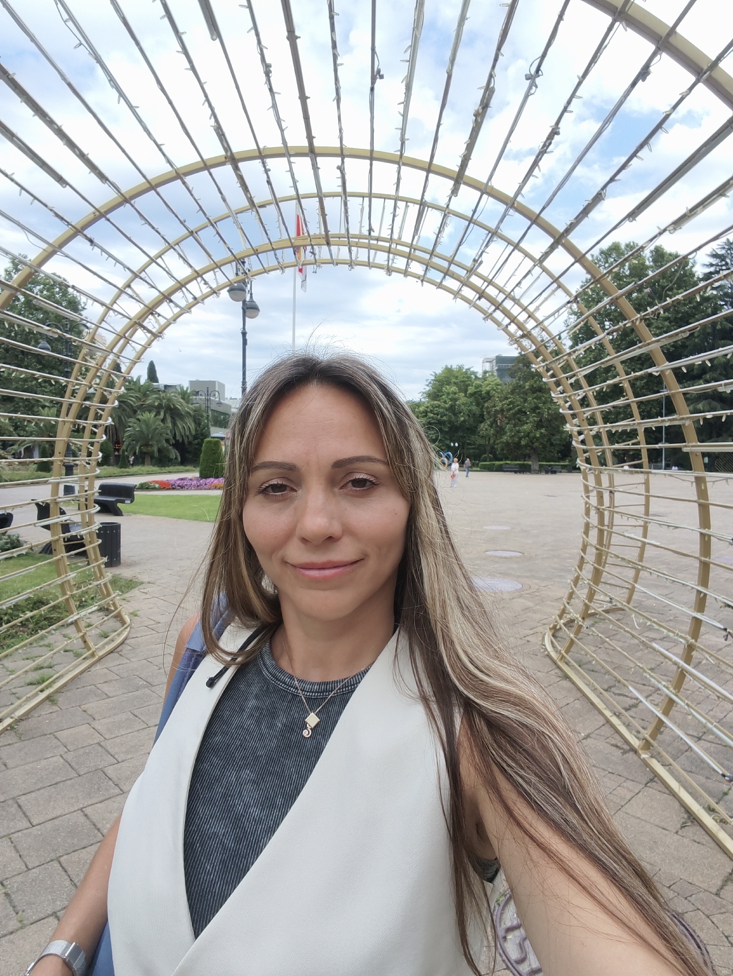 A mid-shot, selfie-style photograph shows a woman with long, wavy, light brown hair and light brown eyes. She is wearing a dark grey shirt with a textured pattern and a white vest. A silver necklace with a small, light-colored pendant is visible. The woman is standing in an outdoor, urban park-like setting, framed by a large, arched, golden structure adorned with small lights. In the background, there are trees, manicured flower beds with purple and pink flowers, a lamppost, park benches, and a trash bin. Further in the background, there are buildings and people walking. The sky above is partly cloudy. This scene is located in the city of Sochi, Russia, likely within a public park or promenade.