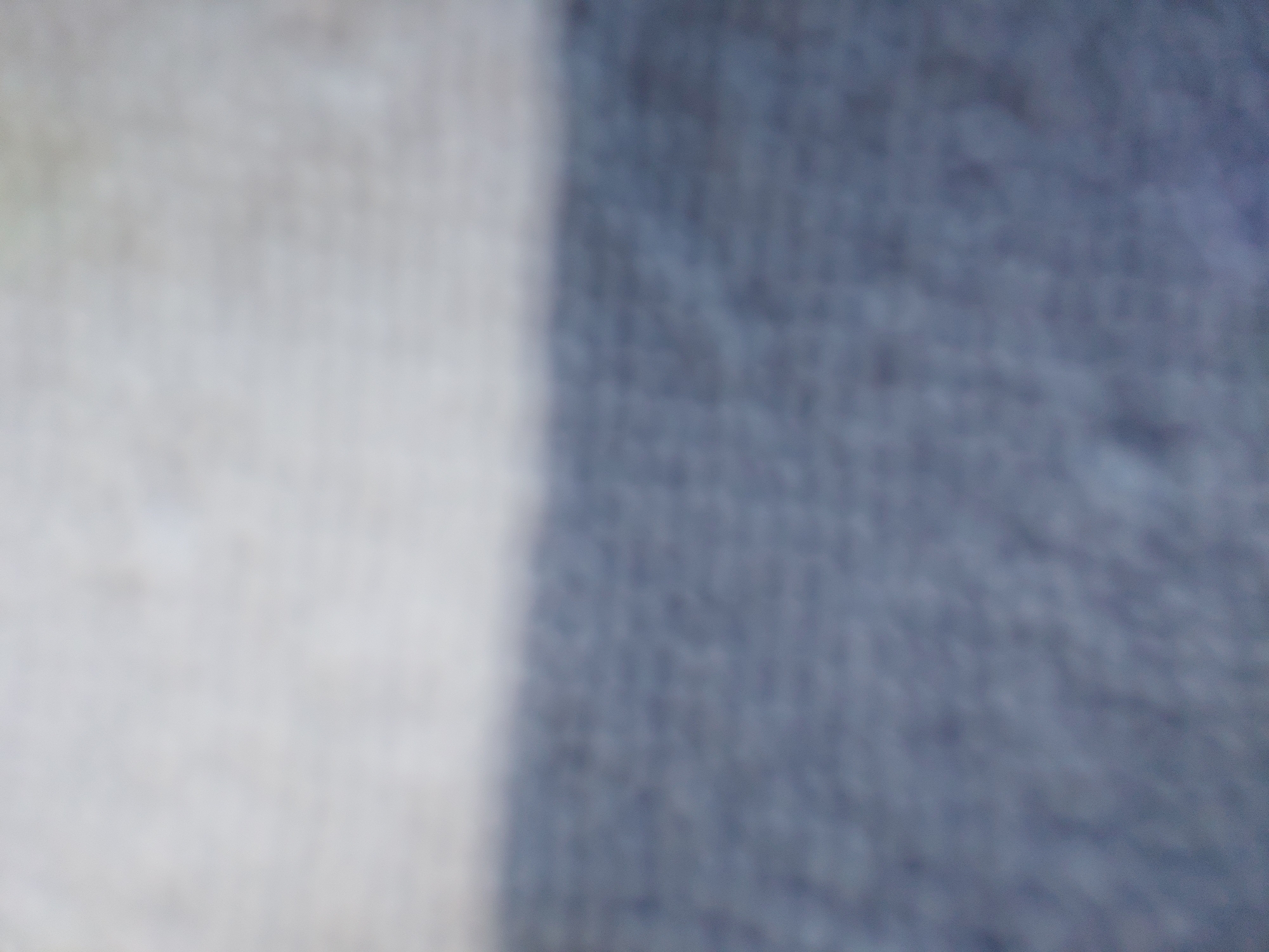 This is a highly blurred, abstract image of a fabric surface. The top portion of the image is a light, off-white color, while the bottom portion is a darker, desaturated blue-gray. The two colors meet in a horizontal line across the middle of the frame. The texture of the fabric appears to be woven or knitted, with small, repetitive patterns visible beneath the blur. There are no discernible objects, people, or environmental features present. The image is too abstract to provide any specific details about a location in Baan Mae Nam, Thailand.
