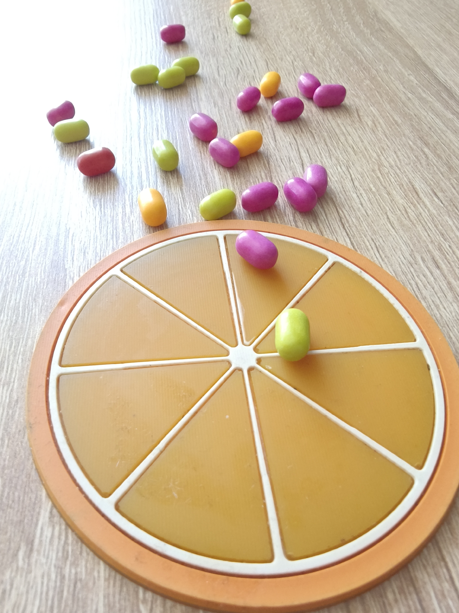 On a light-colored wooden surface, an orange-shaped game board with eight white sections is surrounded by several colorful, bean-shaped candies. One green candy rests within a section on the game board, while a purple candy sits on the board's edge. The remaining candies, colored pink, green, red, and yellow, are scattered on the surface around the game board. The location is not identifiable as the city of Kryvyi Rih, Ukraine.