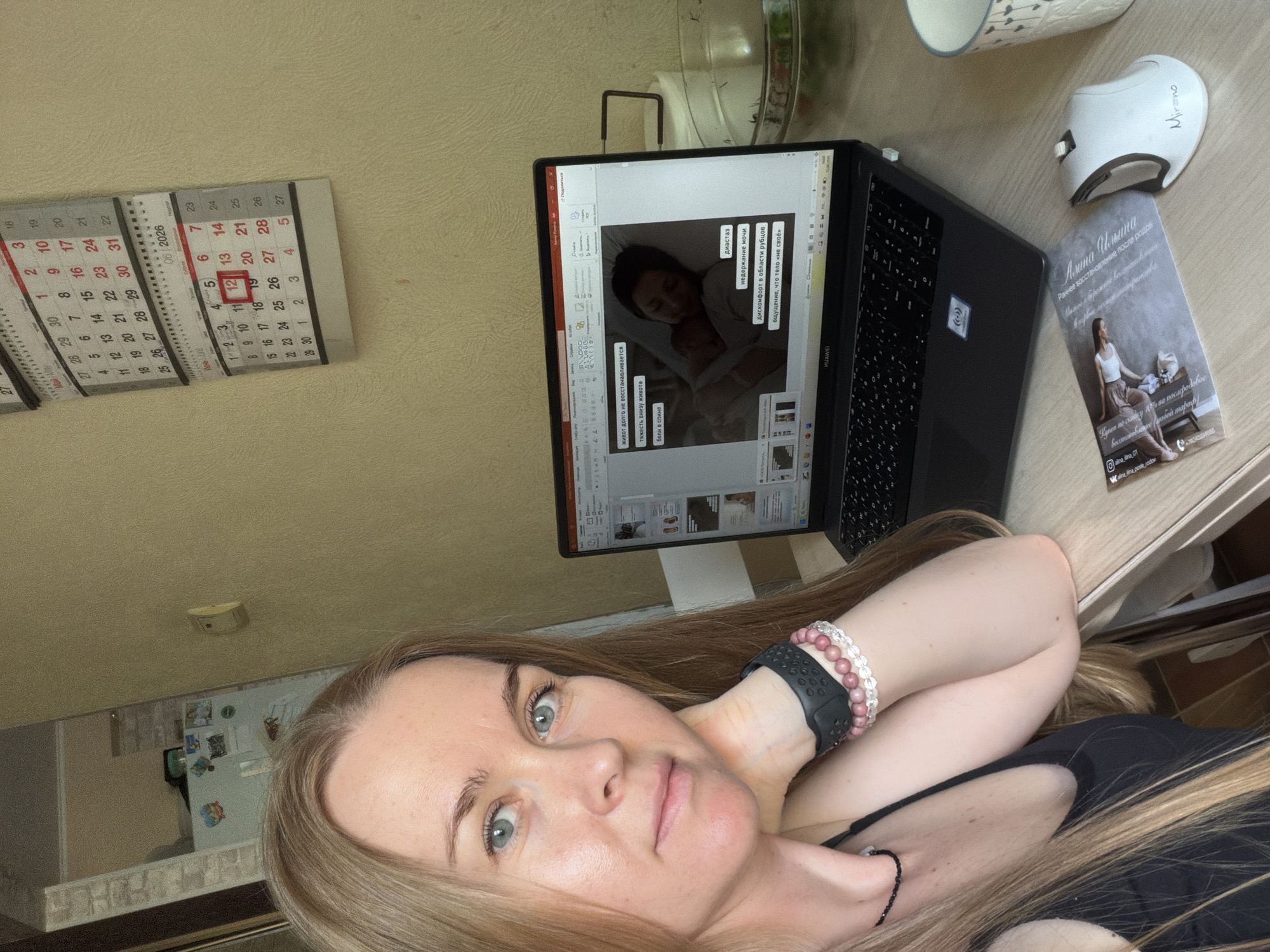 The image shows a woman with long blonde hair sitting at a table in what appears to be a home environment. She is looking towards the camera with a thoughtful expression, resting her chin on her hand. She is wearing a black tank top and several bracelets on her left wrist. In front of her is an open laptop displaying a presentation with images and text. A white computer mouse is to the right of the laptop. A printed card is also visible on the table. A wall calendar with dates for May and June 2026 is hanging on the wall behind the woman. The calendar shows the date "12" circled in red. The room has neutral-colored walls and some kitchen appliances are visible in the background. The scene suggests the woman is engaged in online work or study. The presence of the calendar from 2026 indicates the image was taken in the future, or it is a decorative calendar. The user prompt states the image is from Khabarovsk, Russia, but there are no visual cues in the image to confirm this location.