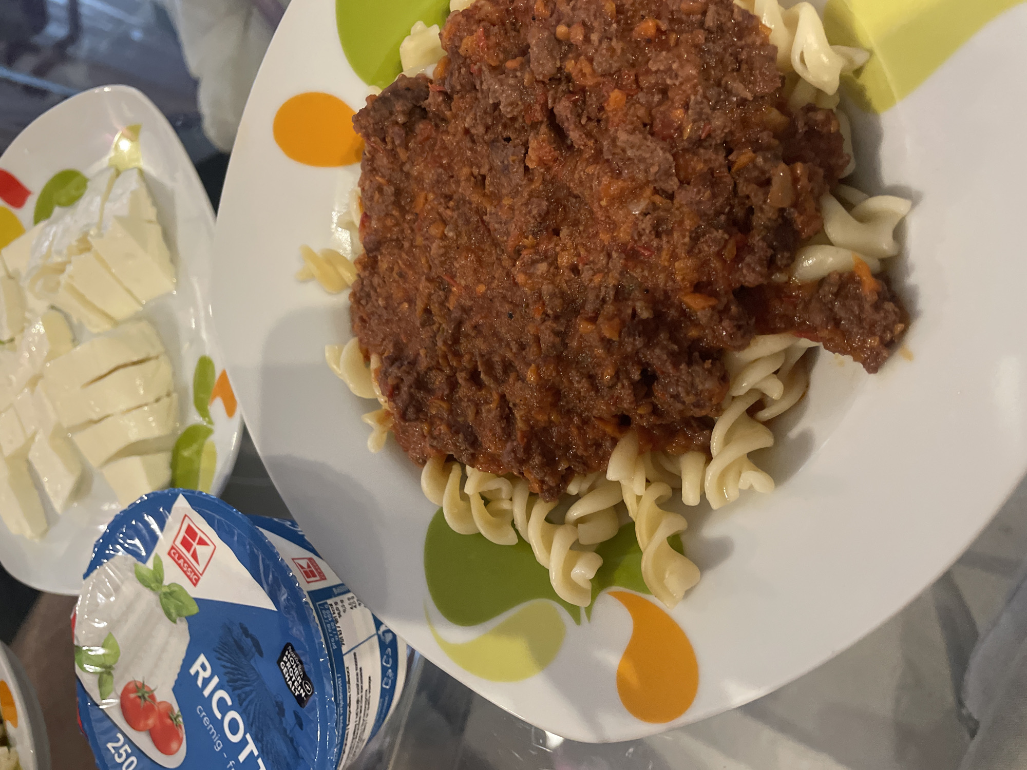 The image shows a plate of rotini pasta with a thick, dark brown sauce, likely a meat sauce, taking up the central portion of the plate. The pasta surrounds the sauce. The plate is white with orange and green circular designs. To the left of the pasta is a plate with cubed cheese and a container of Classic Ricotta cheese. The image was taken in an indoor environment. There is no information provided in the image to place the location in Sofia, Bulgaria.