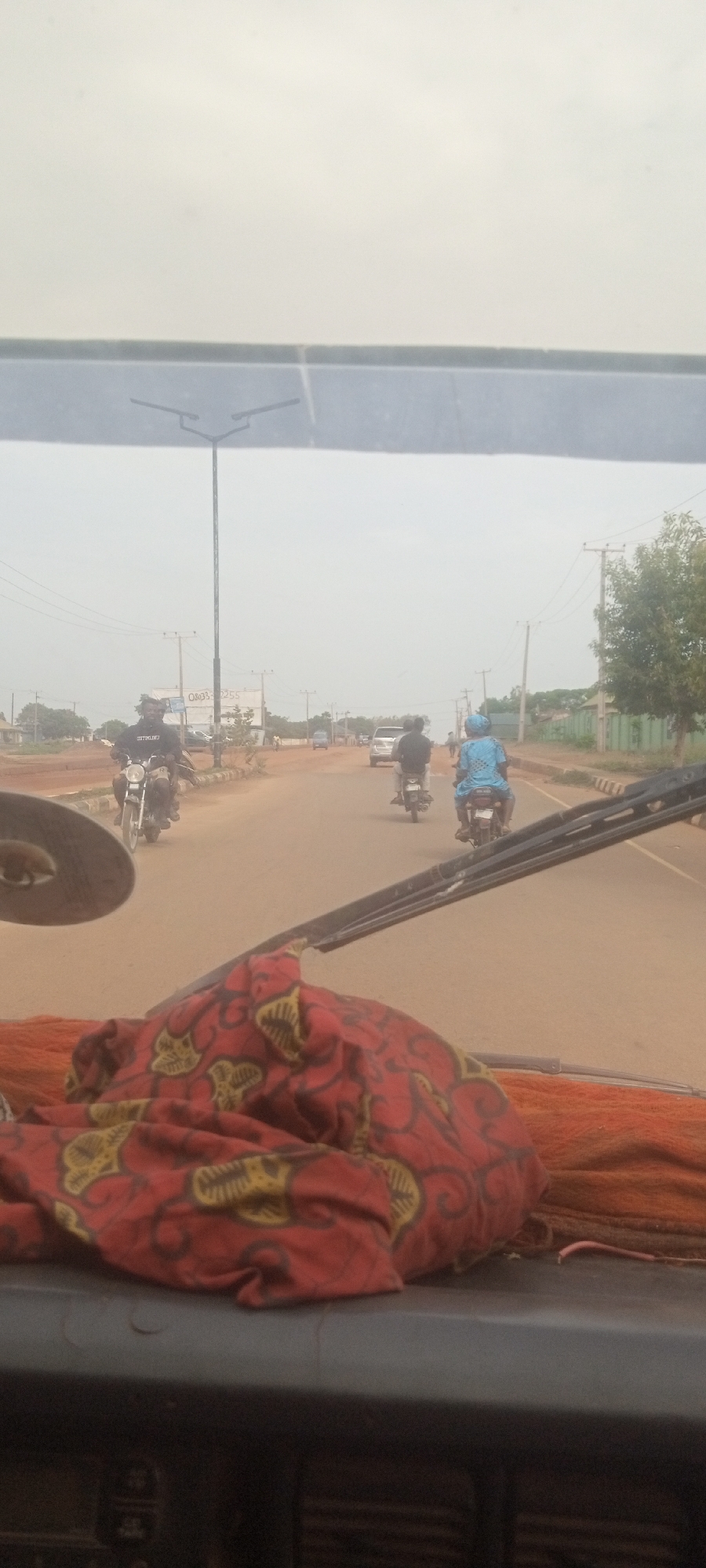 The image depicts a street scene in Television, Nigeria, as viewed from inside a vehicle. The road is dusty and lined with power poles. There are three motorcycles in the distance, two of which are occupied. A light pole is visible on the left side of the road. In the foreground, a patterned red cloth sits on the dashboard of the vehicle, obscuring a clear view.