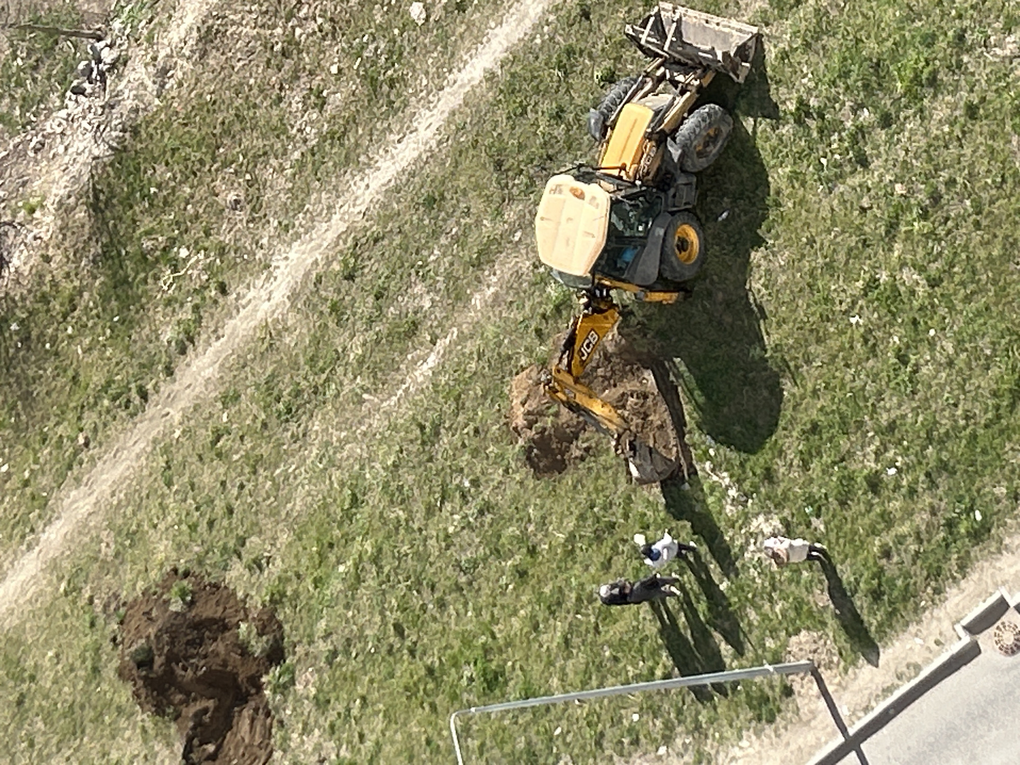 An aerial view captures an outdoor scene in Novoe Devyatkino, Russia, depicting a yellow JCB backhoe loader engaged in ground work on a grassy terrain. The heavy machinery, positioned roughly in the upper center, has its rear excavator arm extended, with a bucket near a fresh pile of dark brown excavated earth. Another, smaller excavation or pile of earth is visible further to the left. The backhoe also features a front loader bucket.
Three individuals are standing on the grass near the machinery. One person in a light top and dark pants is closer to the excavator arm, while another, possibly wearing a cap, stands a little further. A third person in light attire and dark pants is positioned closer to the foreground, near a metal railing structure. Their long shadows indicate it is daytime, likely mid-morning or afternoon, under clear skies.
The ground is predominantly green grass, interspersed with worn dirt paths or lighter, less vegetated areas. In the foreground, a gray metal railing runs horizontally, and to the far right, a section of paved sidewalk with a visible manhole cover suggests proximity to developed infrastructure. The overall scene suggests a construction or maintenance operation taking place in a grassy, possibly urban fringe or park-like setting. The brand "JCB" is clearly visible on the excavator arm.