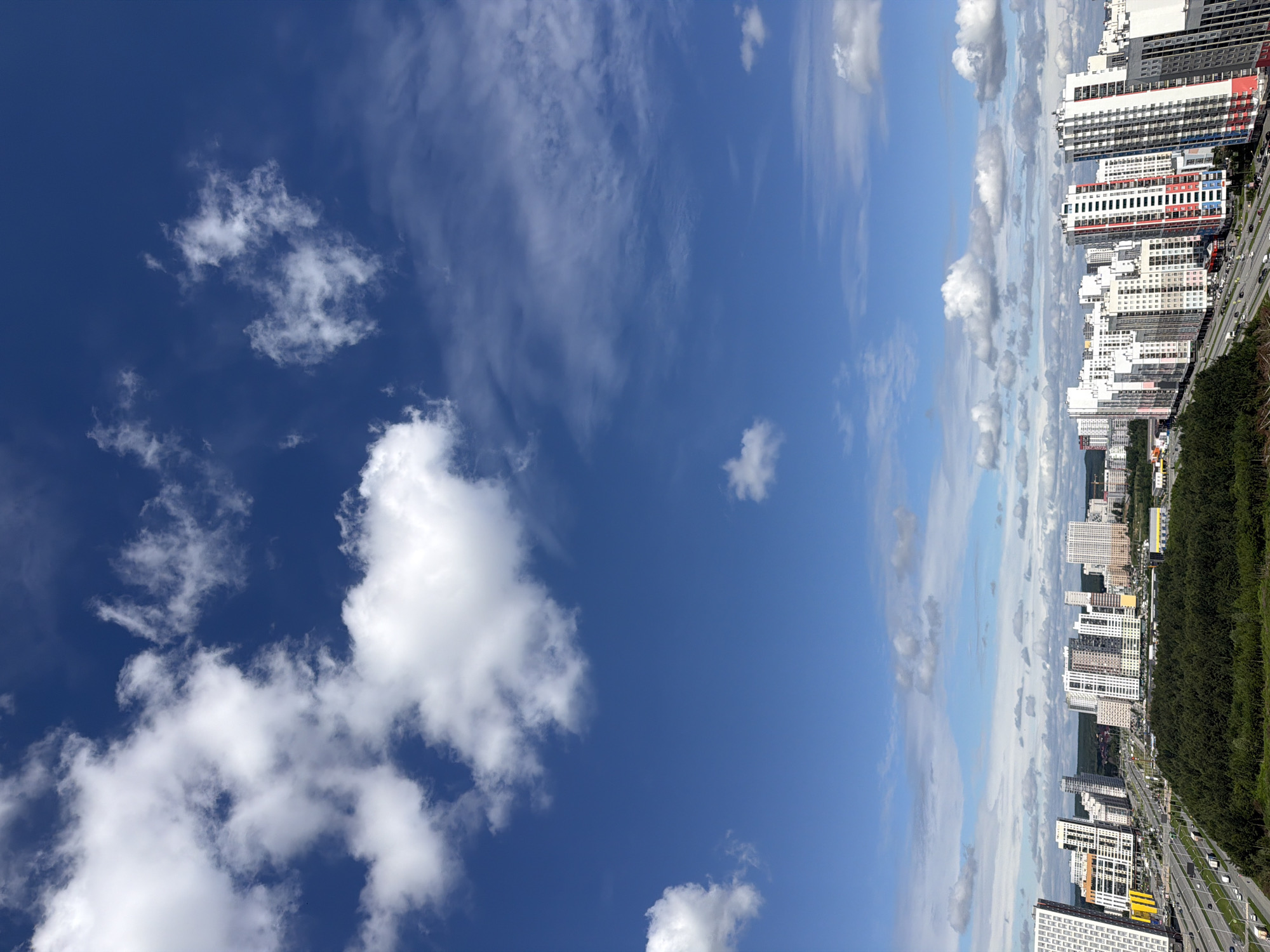 This is an aerial, vertical, daytime photograph of a modern urban cityscape under a bright blue sky with scattered white cumulus clouds. The scene is dominated by a dense collection of residential buildings of varying heights and architectural styles, many featuring colorful facades. A multi-lane highway runs through the foreground, with a few vehicles visible. A large expanse of dark green trees separates the highway from the buildings. The city appears to be located in a relatively flat area.