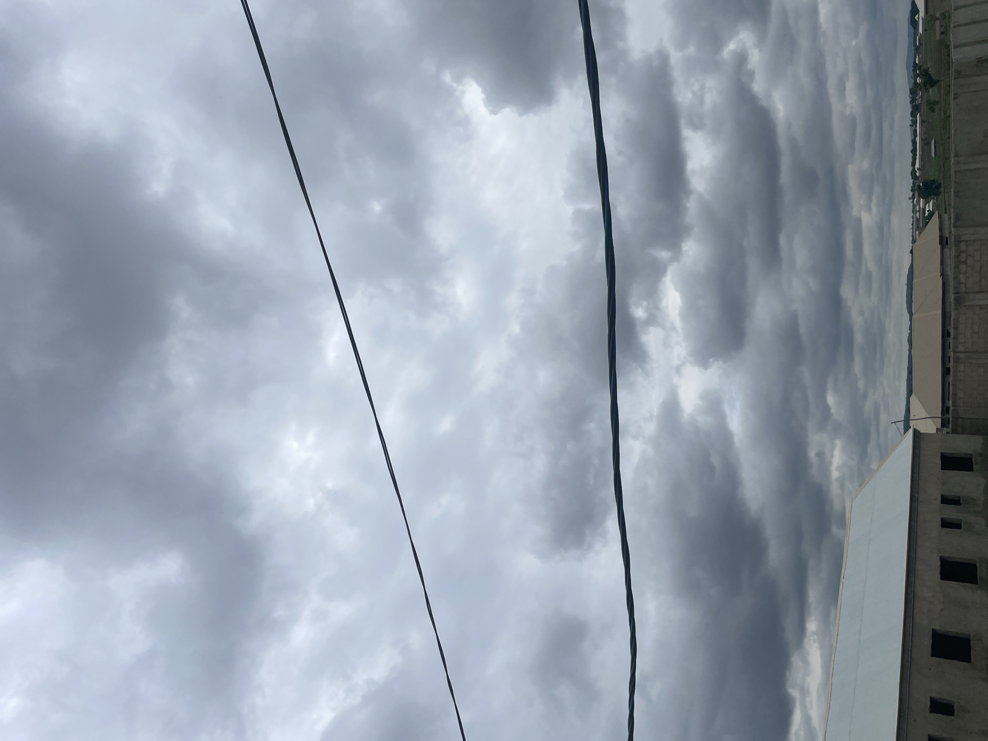 The image captures a cloudy, overcast sky above a developing urban or suburban area, possibly in Barako, Nigeria. Two prominent dark power lines stretch horizontally across the frame, dividing the sky. Below the sky, a partially constructed building with a light-colored roof and dark, unglazed window openings occupies the lower left portion of the image. In the background, more buildings with similar roofing materials are visible, suggesting ongoing construction or a growing community. The landscape appears relatively flat, with some distant hills or elevated terrain. The overall impression is one of an active construction site under a moody, cloud-filled sky.