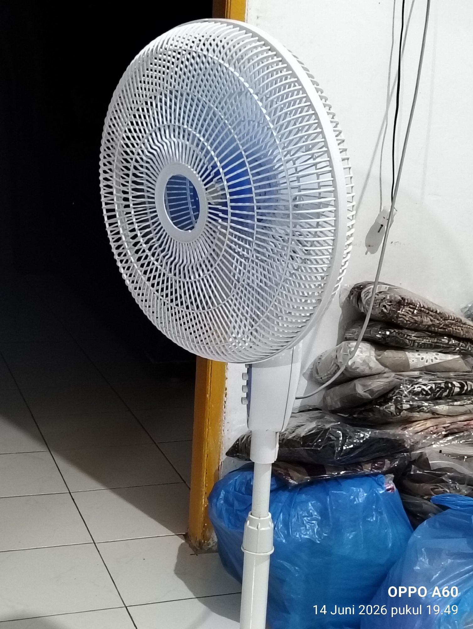 A white standing fan is positioned in the foreground, angled towards the left, obscuring a portion of the background. The fan's blades are visible through its white grill. To the right of the fan, a stack of items wrapped in clear plastic and patterned fabrics is visible. Below these items, two large blue plastic bags are prominent. The floor is tiled with white squares and grout lines. A yellow-painted door frame is visible on the right edge of the image. The overall lighting suggests an indoor environment, possibly a room. The bottom right corner displays text: "OPPO A60" and "14 Juni 2026 pukul 19.49". The location is not specified in the image.