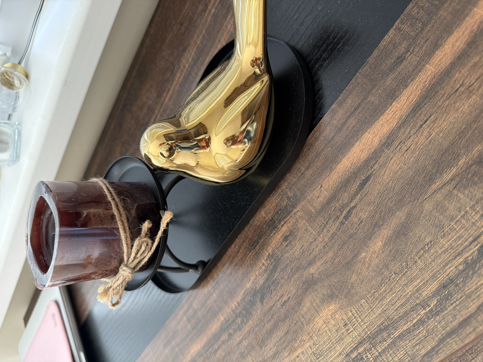 The image shows a close-up view of a wooden surface with a black tray containing a dark brown square candle on a black candle holder, and a shiny gold bird figurine. The candle is tied with twine. The scene is indoors, with a white windowsill visible in the background, reflecting some objects. The wooden surface has a dark brown, textured grain. There are no people present in the image, and no identifiable landmarks or features specific to Pazardzhik, Bulgaria.